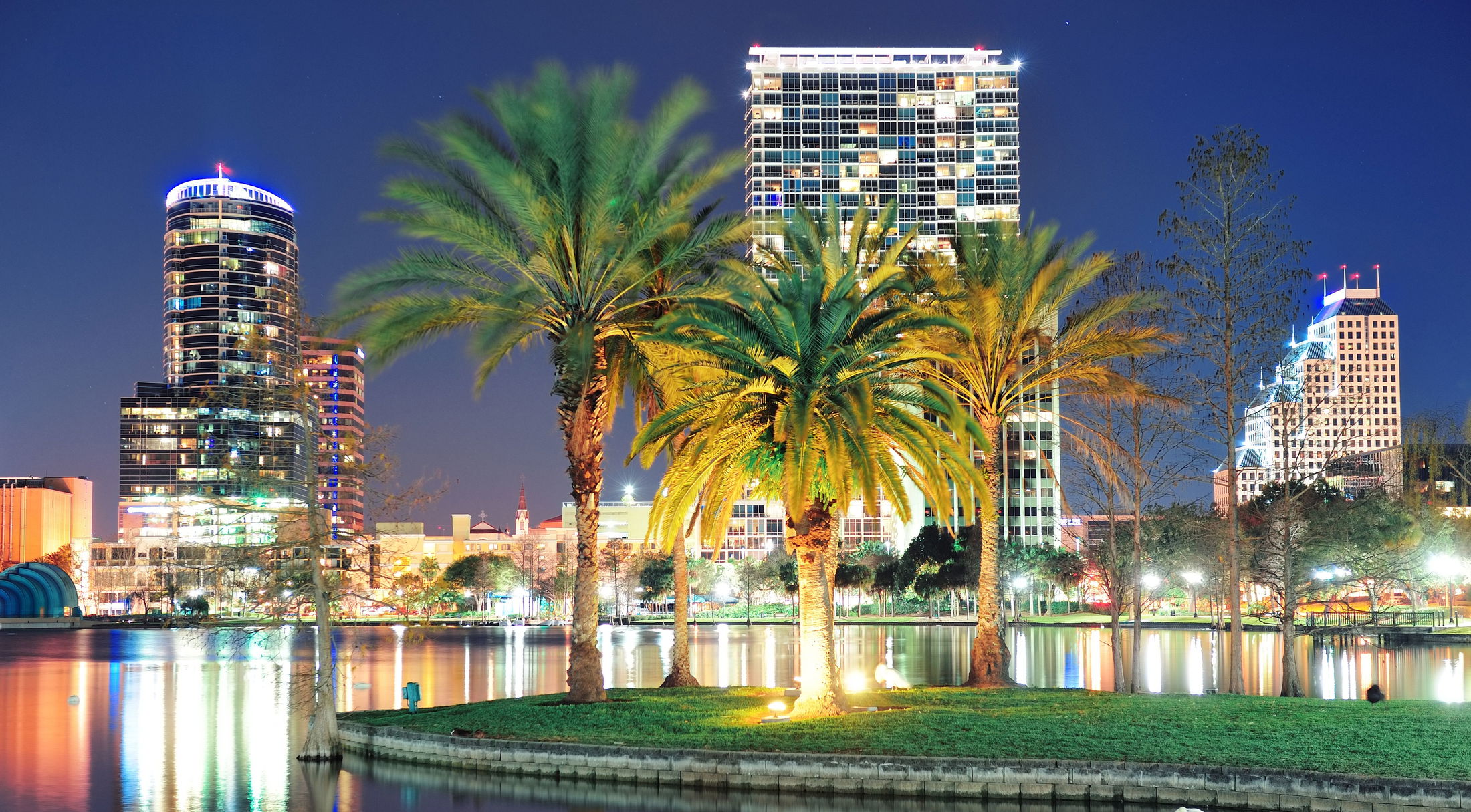 Vibrant nighttime view of downtown Orlando, Florida, showcasing illuminated skyscrapers and palm trees reflecting on Lake Eola.
