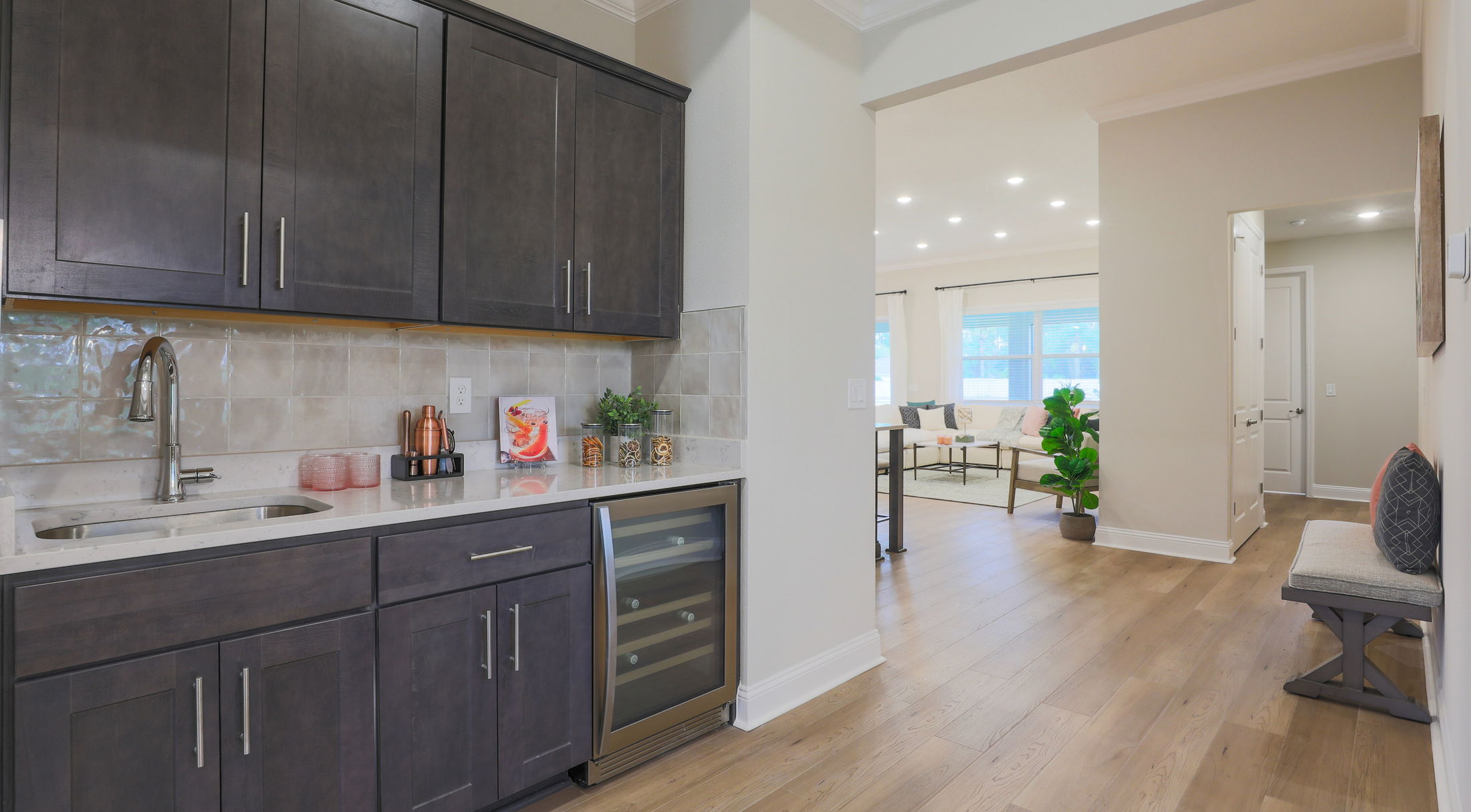 Modern kitchen with dark cabinets, marble countertop, and wine cooler, overlooking a bright, open living room with hardwood floors.