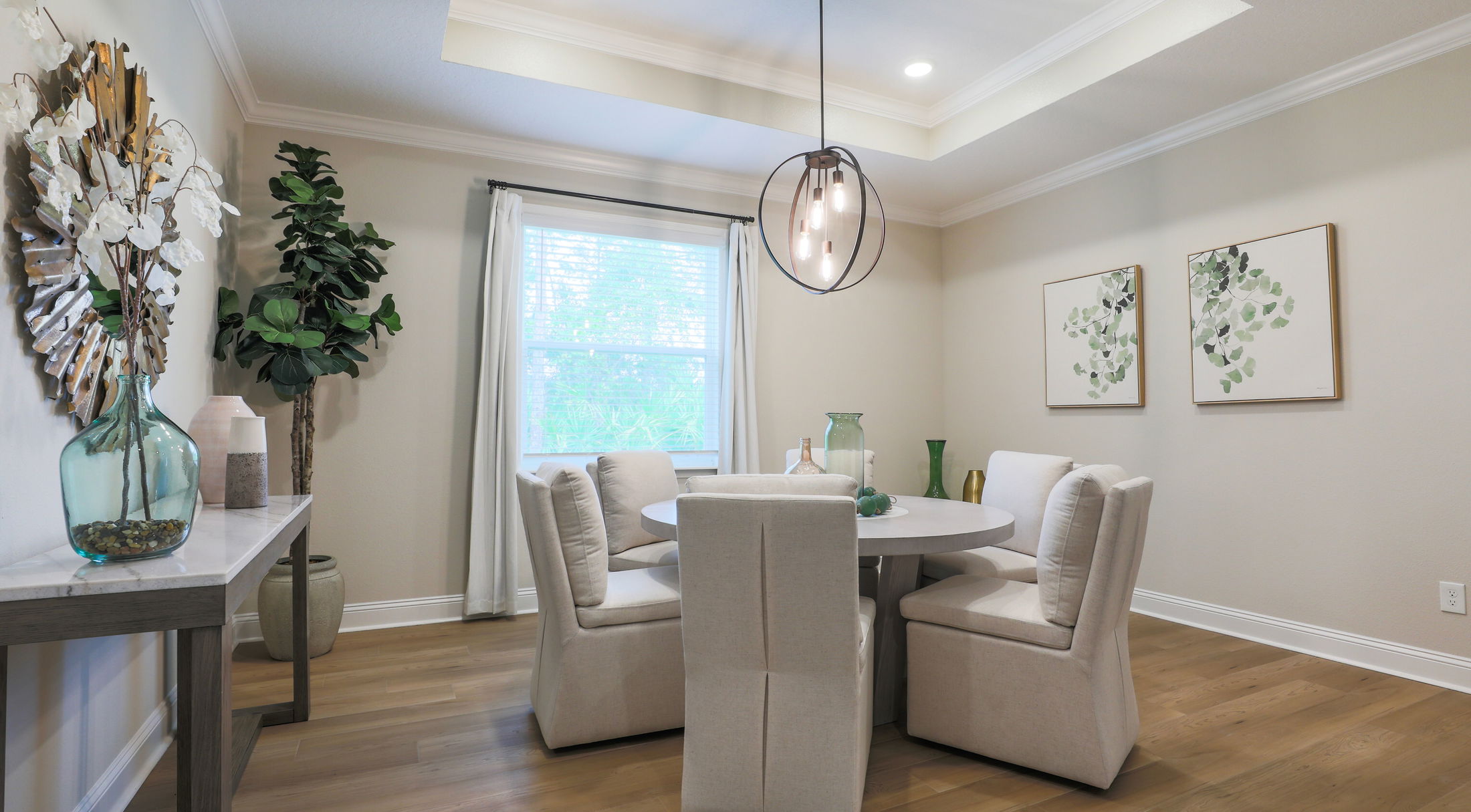 Elegant dining room with modern chandelier, round table, upholstered chairs, and bright decor featuring plants and wall art.