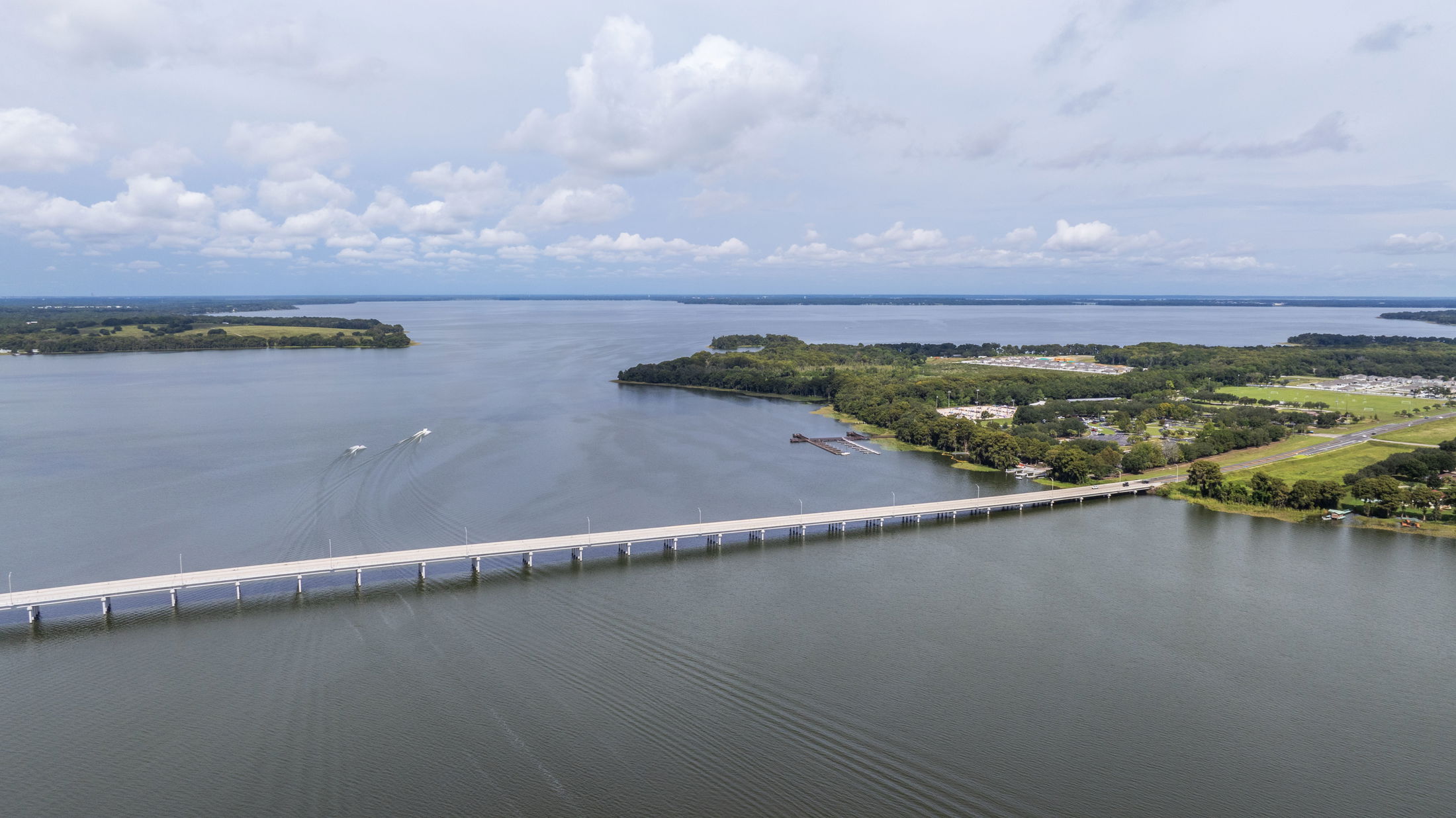 Aerial view of a long bridge spanning across a calm lake with surrounding lush green landscape and two boats leaving trails in the water under a partially cloudy sky.