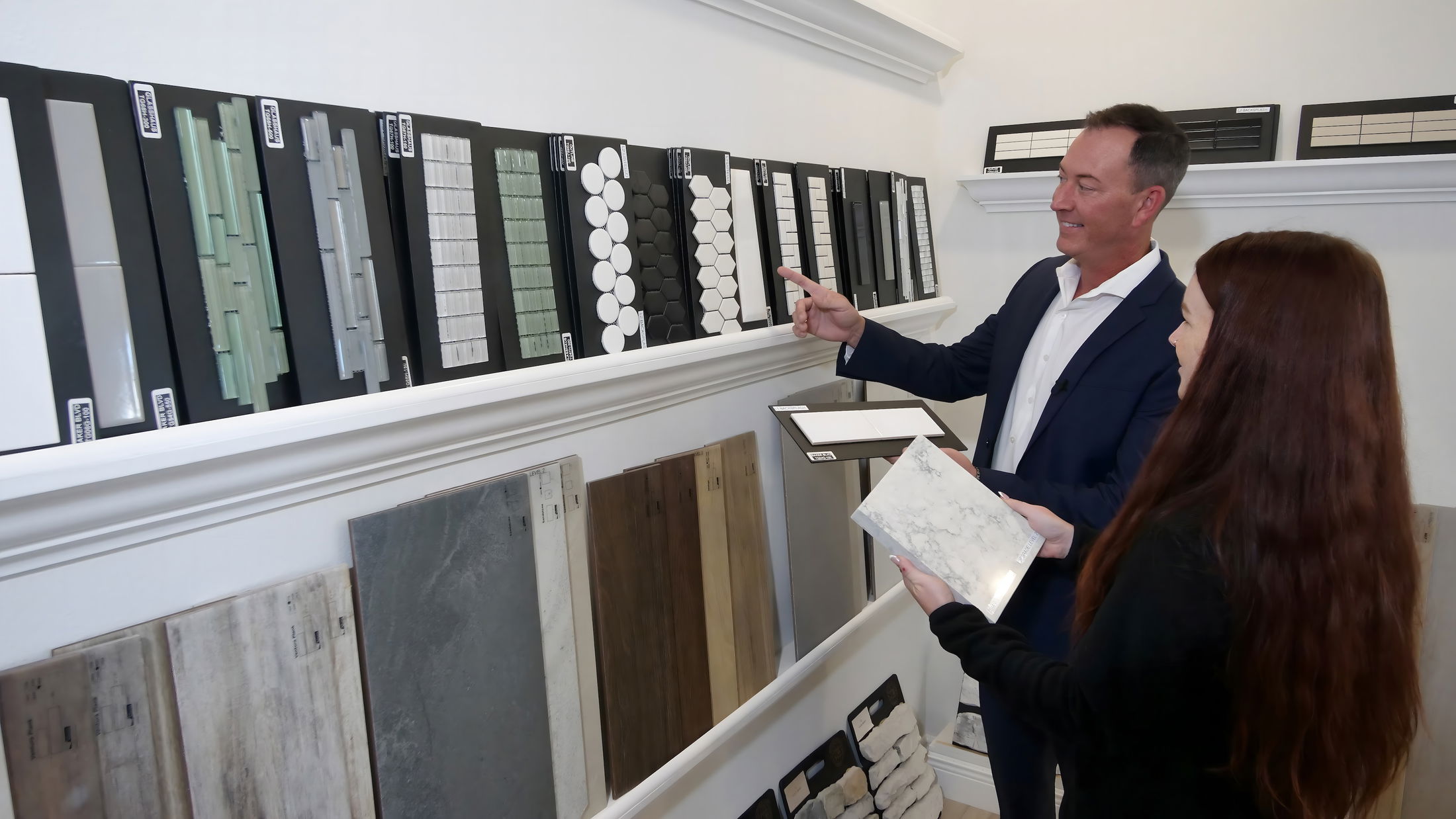 A professional interior designer assists a client in selecting various tile samples from a display wall in a modern design studio.