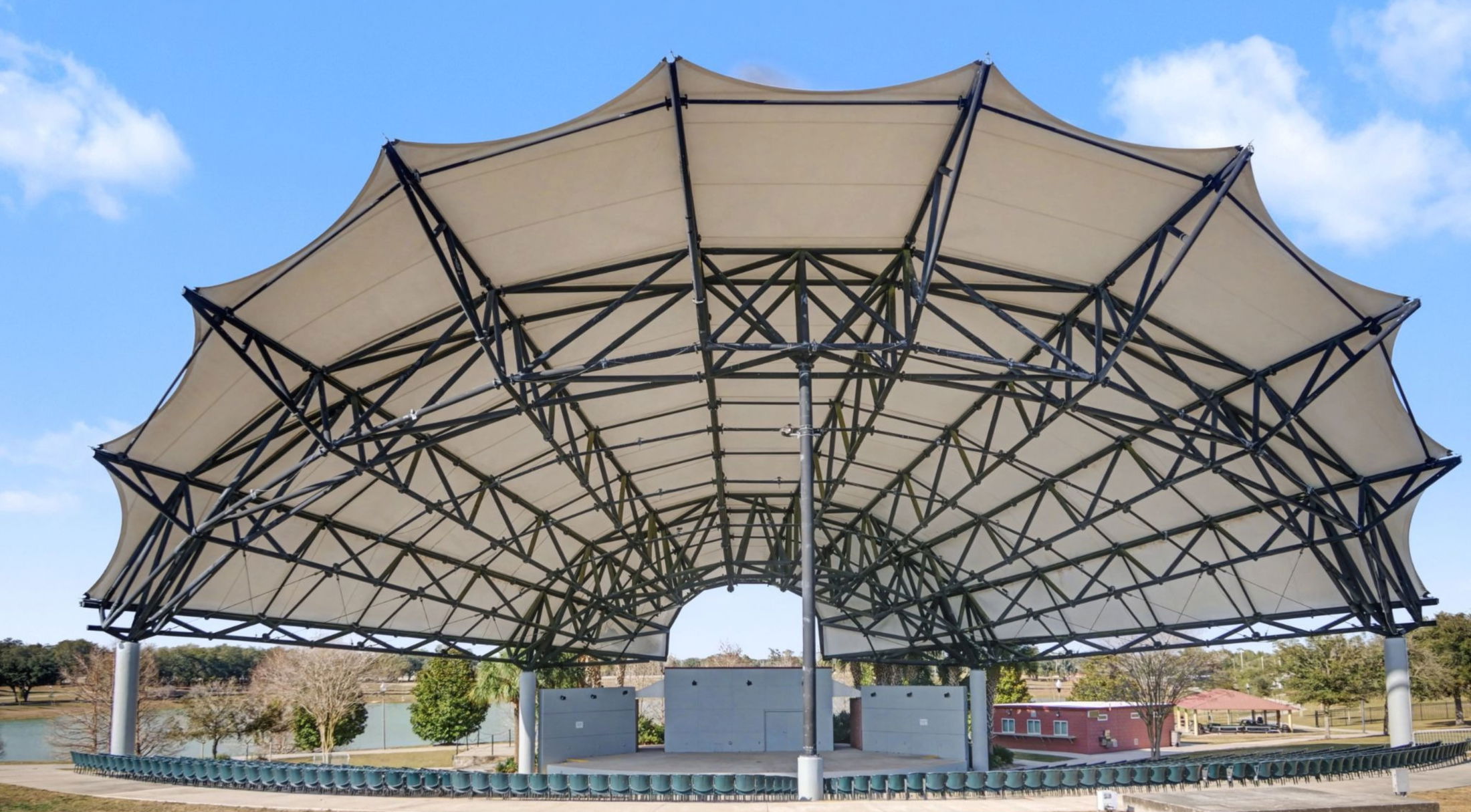 A large, modern outdoor concert pavilion with a distinctive canopy design and surrounding seating located near a lake.