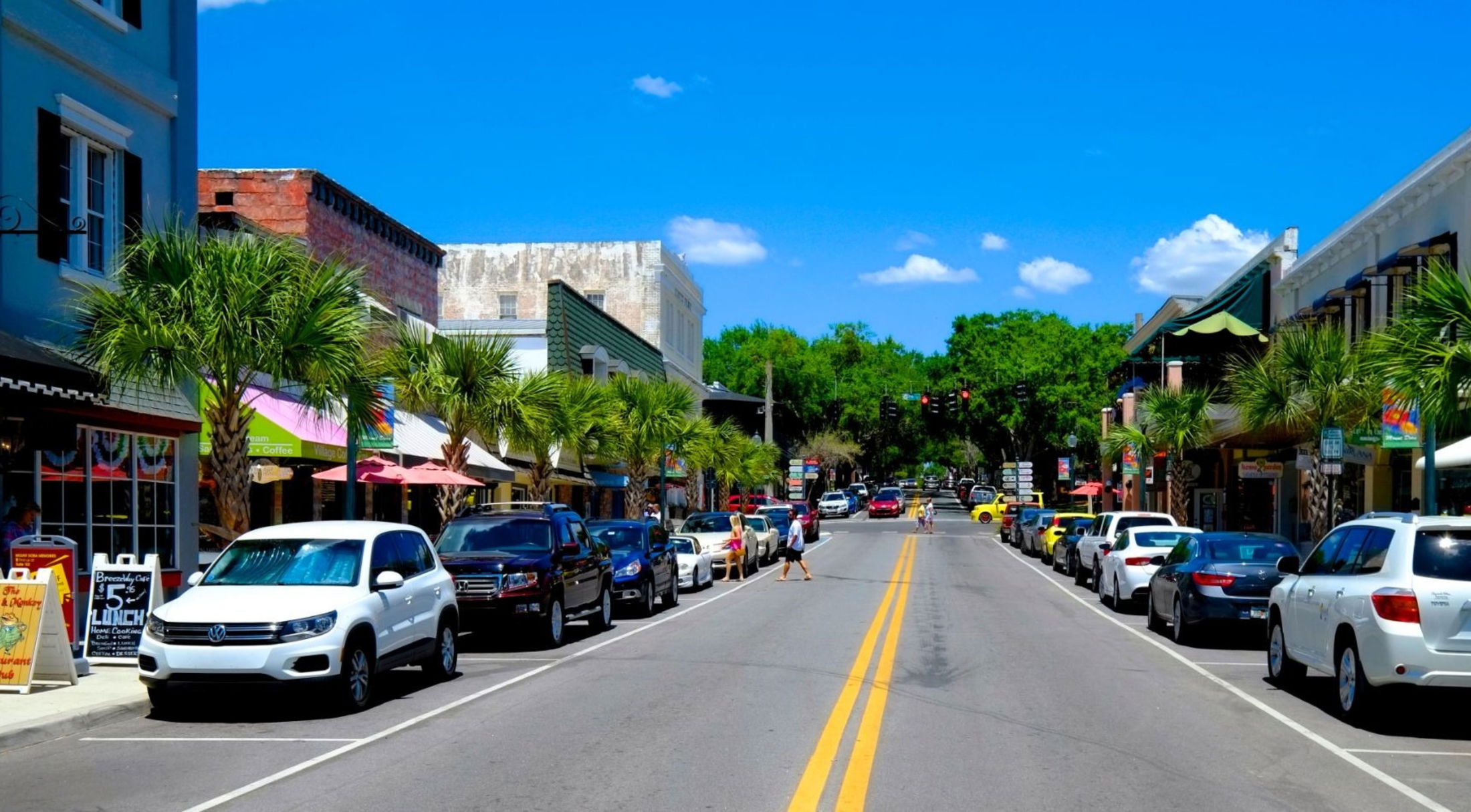 A vibrant street scene in a small town featuring palm trees, parked cars, and shops under a clear blue sky.