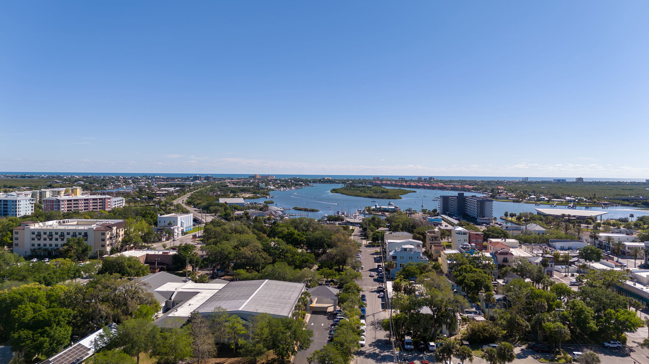 Aerial view of a coastal cityscape with buildings, greenery, and waterways under a clear blue sky.