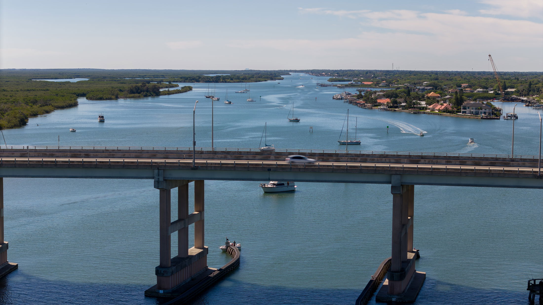 A scenic view of a busy waterway with sailboats and motorboats beneath a concrete bridge, surrounded by lush greenery and coastal homes.