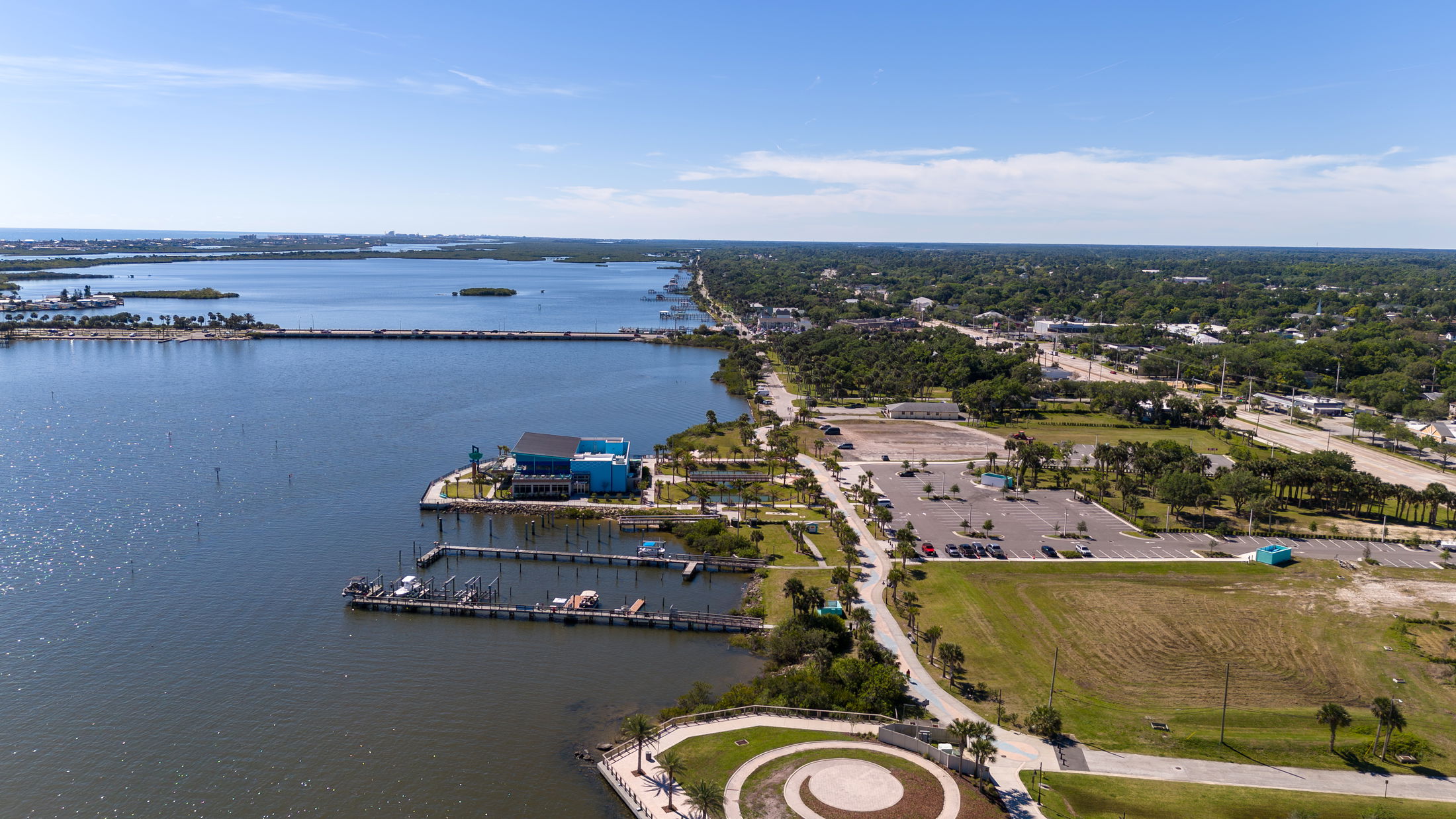 Aerial view of a coastal cityscape with a marina, blue building, and surrounding greenery along a scenic waterfront.