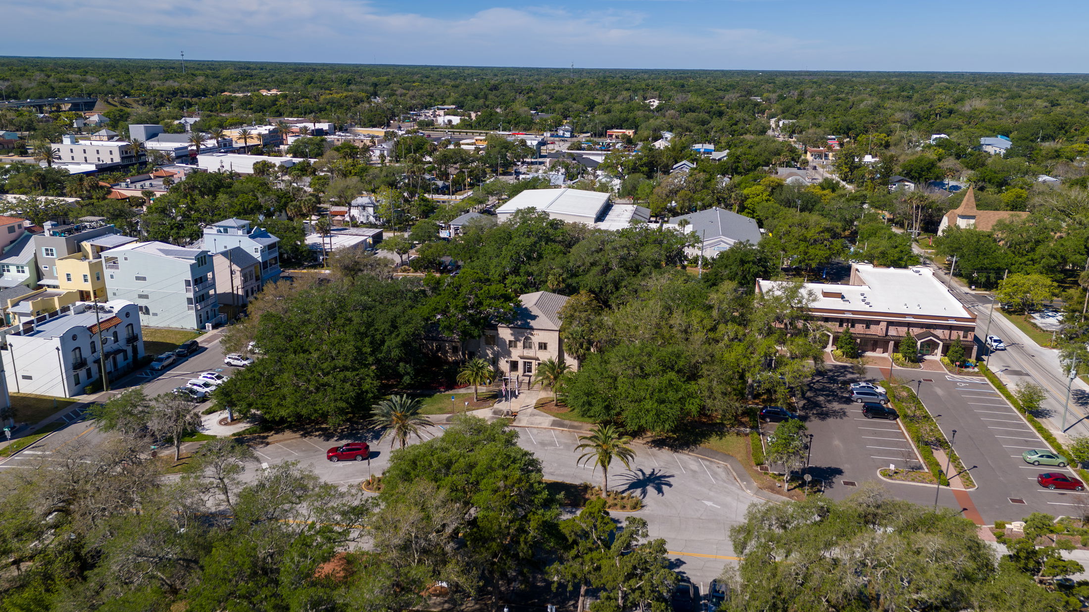 Aerial view of a small town with residential buildings, lush greenery, and parking lots under a clear blue sky.
