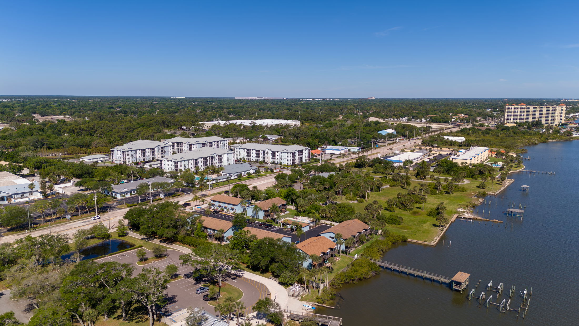 Aerial view of a residential and commercial waterfront area with green spaces and a vibrant coastline under a clear blue sky.