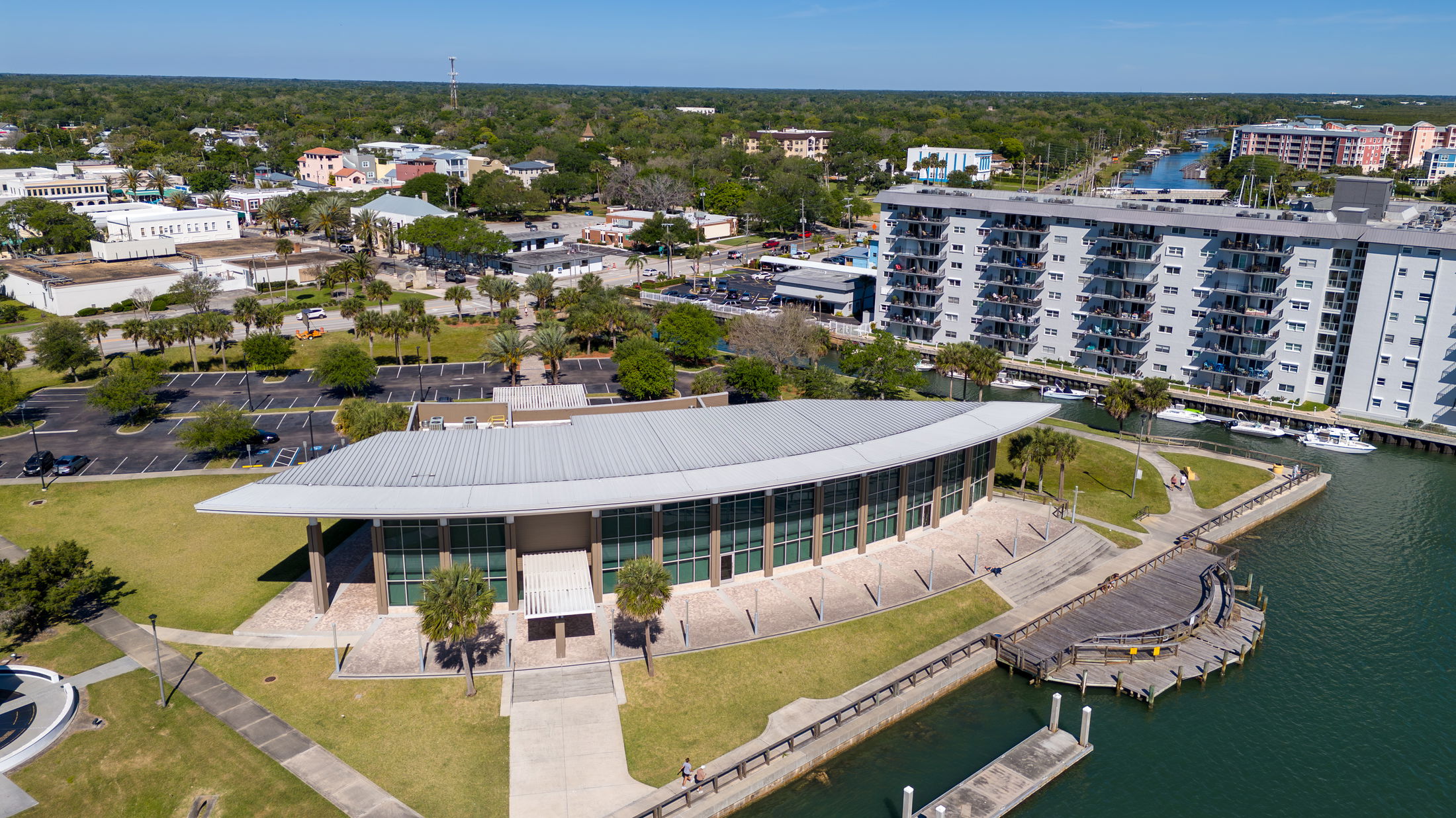 Aerial view of a modern waterfront building with a curved roof, surrounded by parking lots, lush greenery, and a residential area in New Smyrna Beach, Florida.