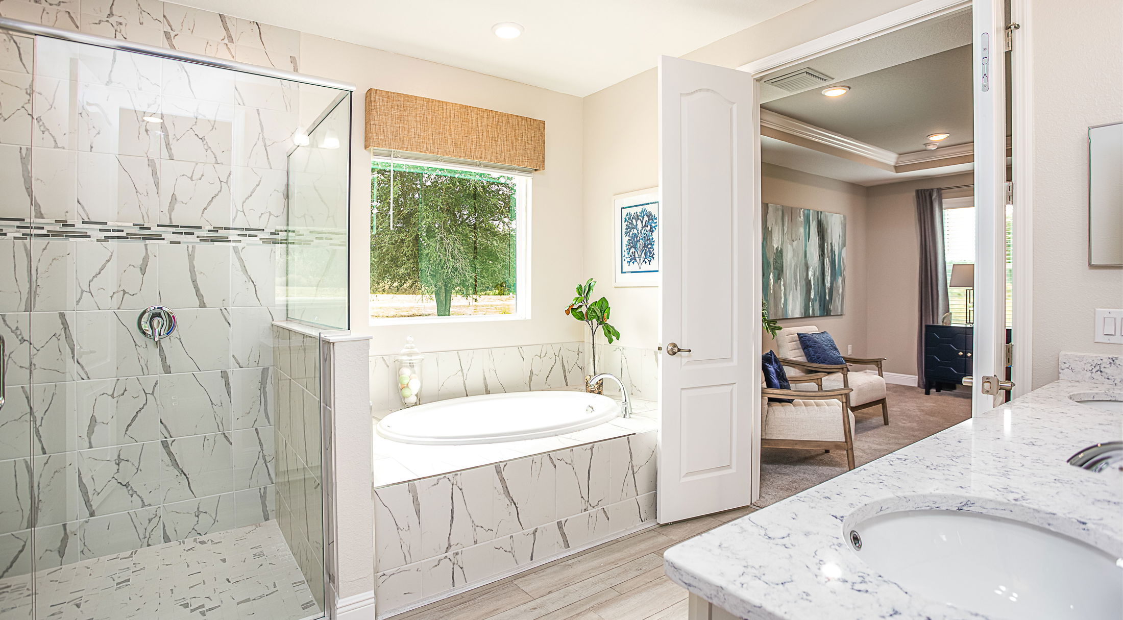 Luxurious modern bathroom with marble-tiled shower, soaking tub, and dual-sink vanity leading into an elegantly decorated bedroom.