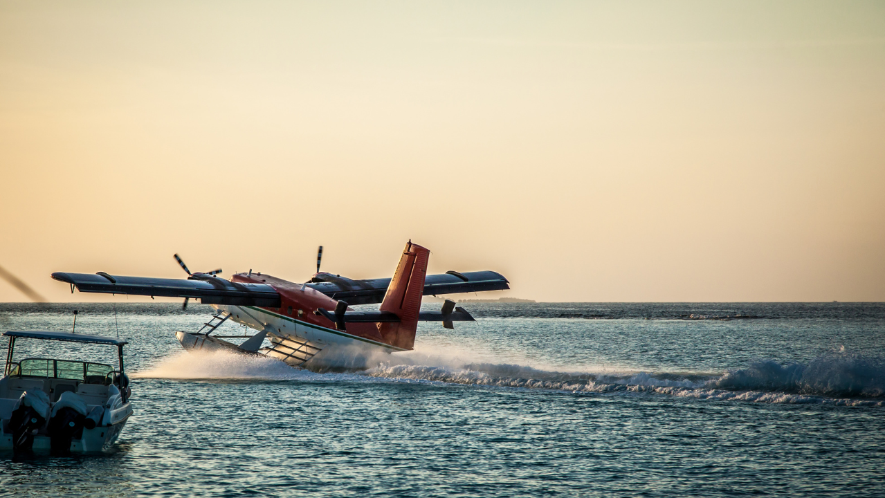 A seaplane with red and white livery takes off from the ocean at sunset as a small motorboat floats nearby.