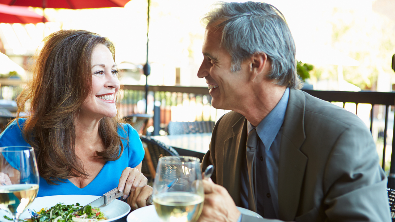 A smiling couple enjoying a meal together at an outdoor restaurant with wine and salads.