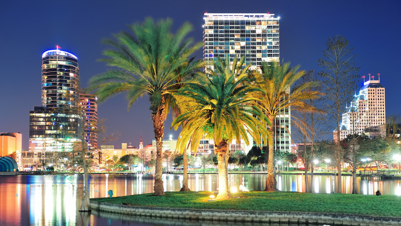Night view of downtown Orlando skyline with illuminated palm trees reflecting in Lake Eola.