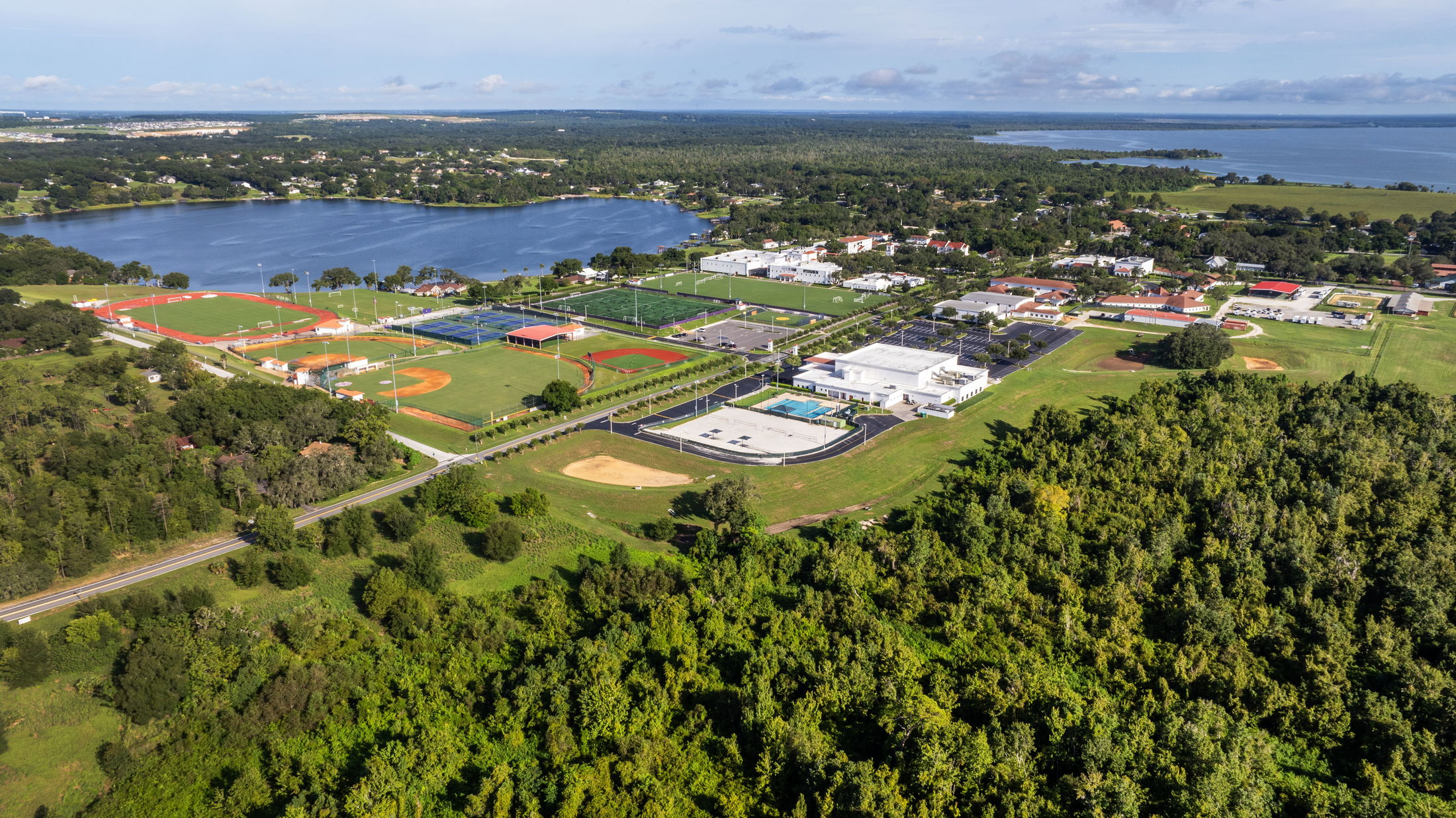 Aerial view of a large educational and sports complex surrounded by lush greenery and a lake, featuring multiple fields, courts, and buildings.
