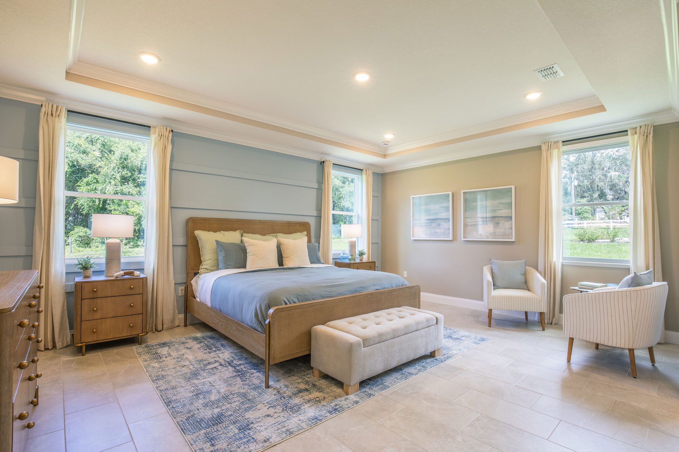 Modern, spacious bedroom with a bed, cushioned bench, twin armchairs, and large windows overlooking greenery.