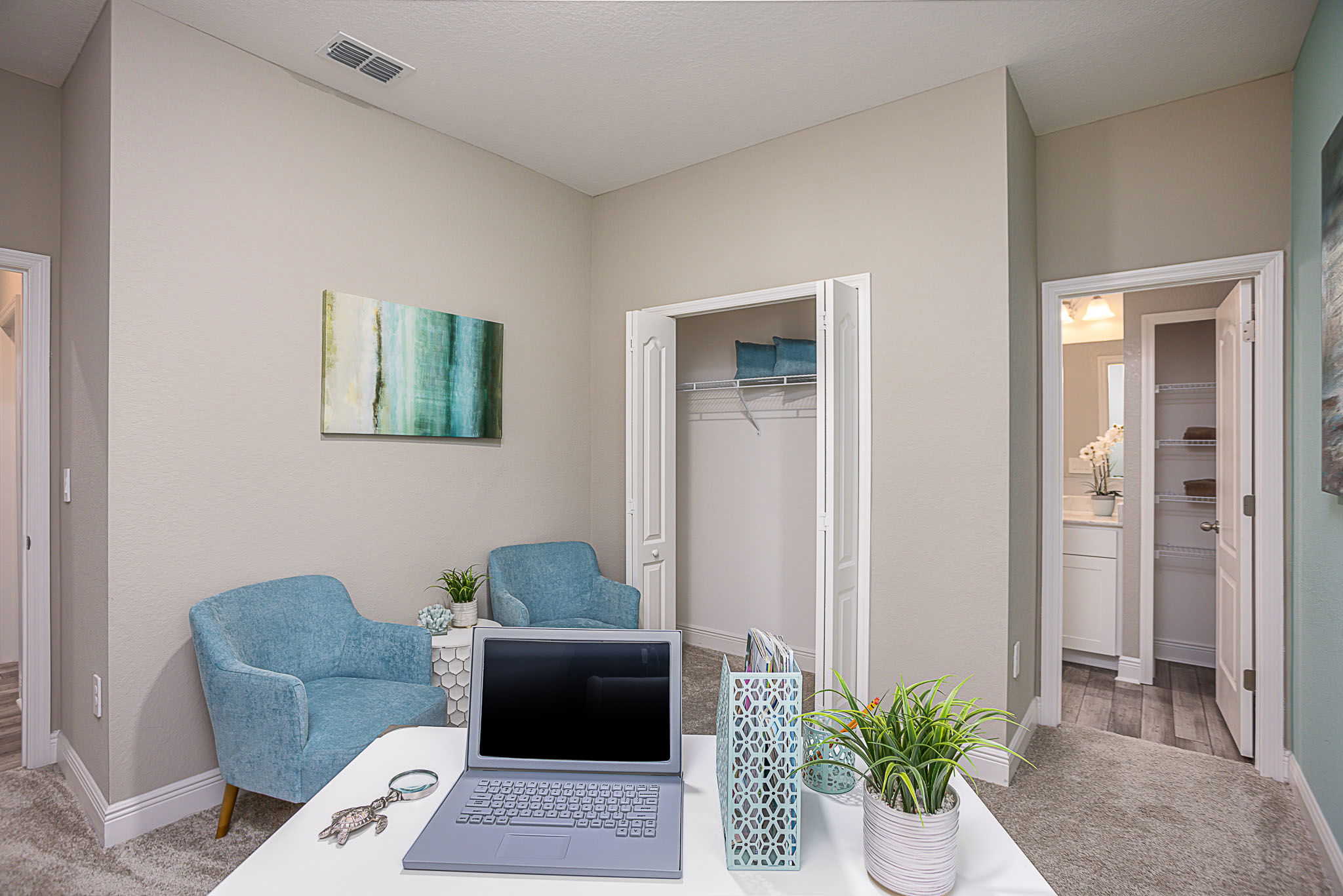 Modern home office setup with a laptop, plants, and decor in a room featuring blue chairs, a closet, and an adjoining bathroom.