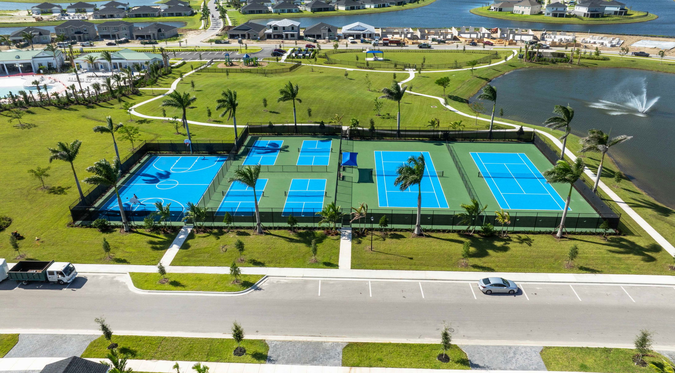 Aerial view of a community sports facility featuring basketball and tennis courts surrounded by green lawns and palm trees.