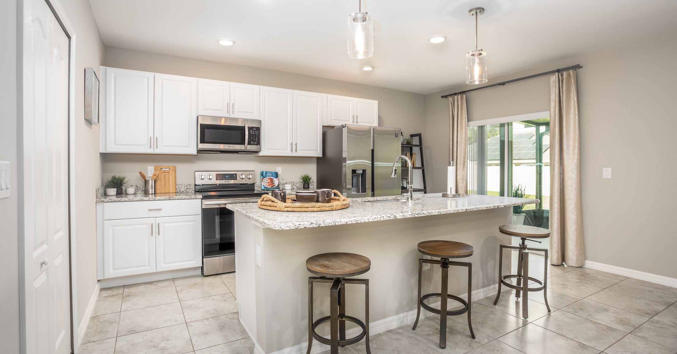Modern kitchen interior with white cabinets, stainless steel appliances, granite countertops, and bar stools, illuminated by pendant lights.
