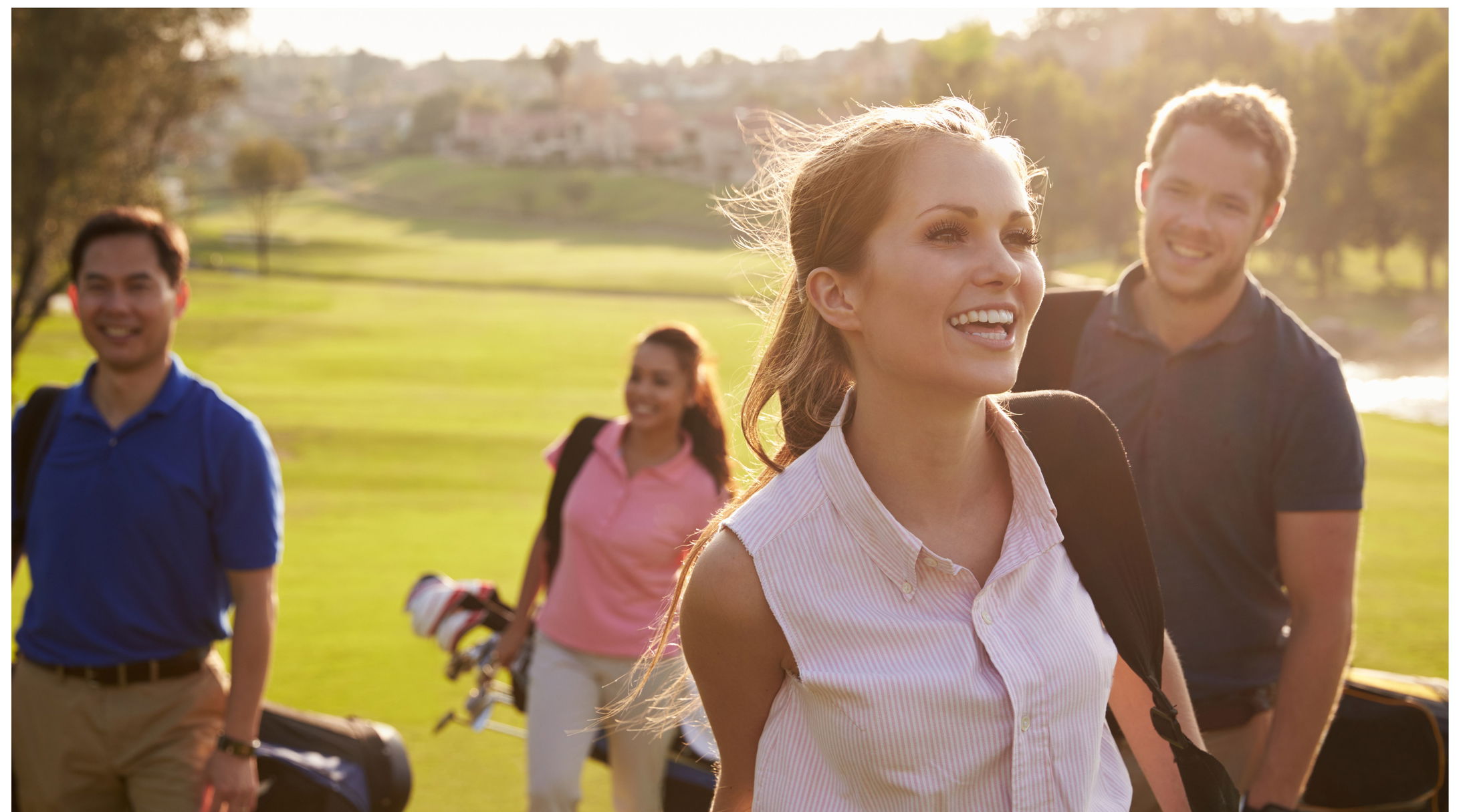 Group of four cheerful friends carrying golf bags and walking on a sunny golf course.