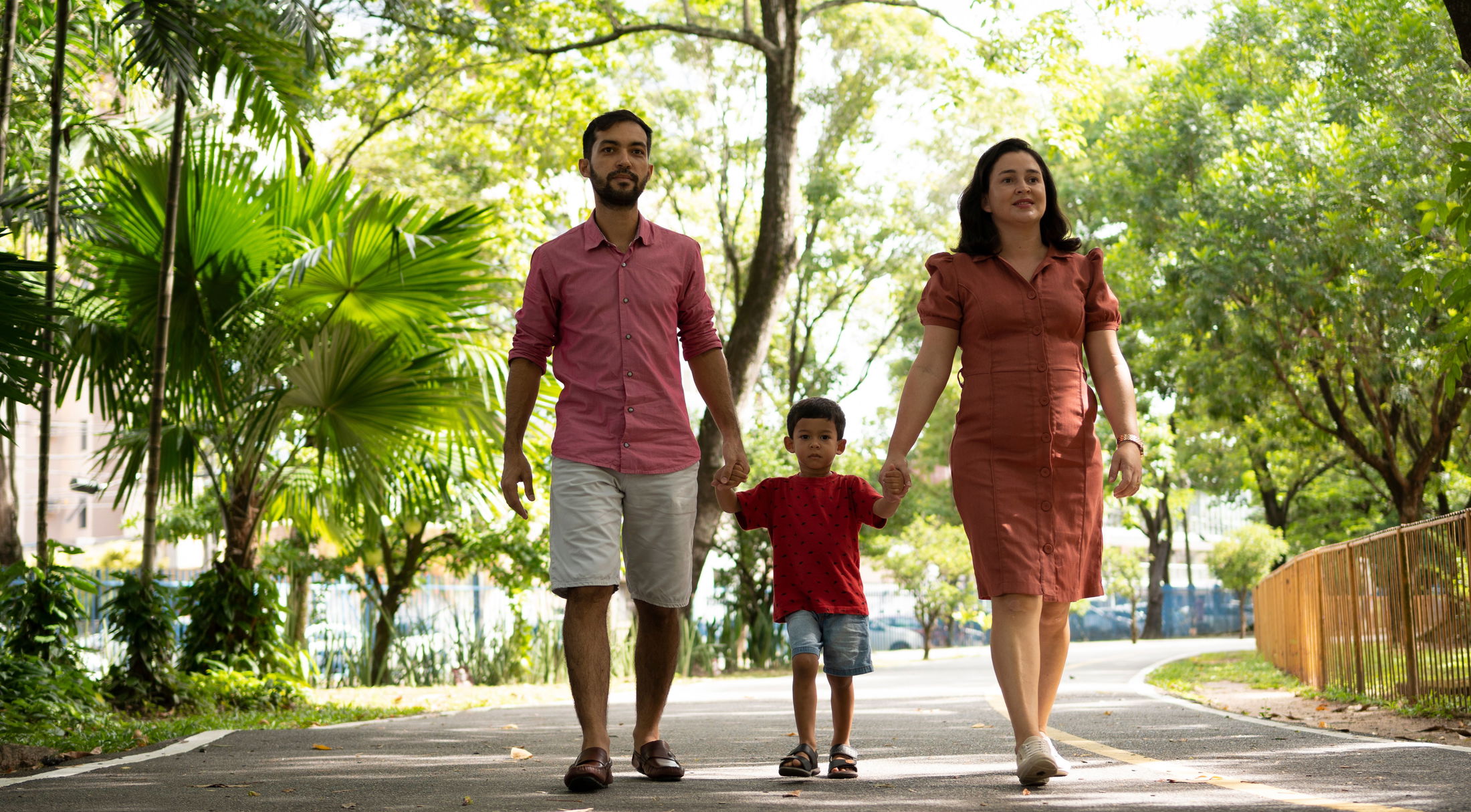 A family of three, holding hands and walking on a tree-lined path in a park on a sunny day.