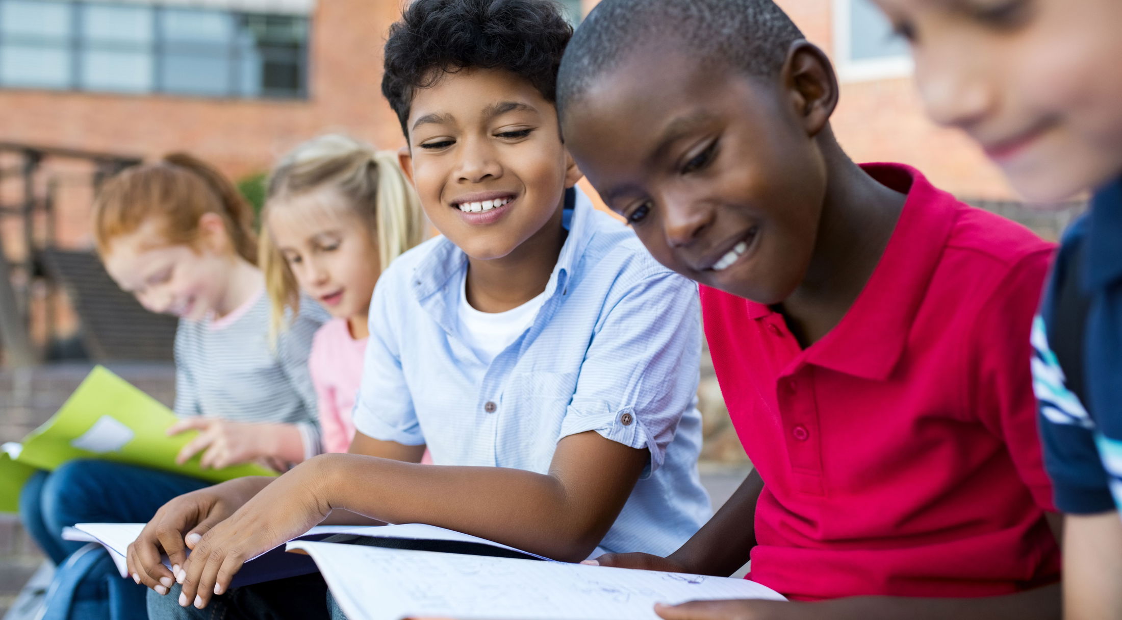 A group of diverse children sitting together outside, reading and smiling, promoting a love for learning and friendship.