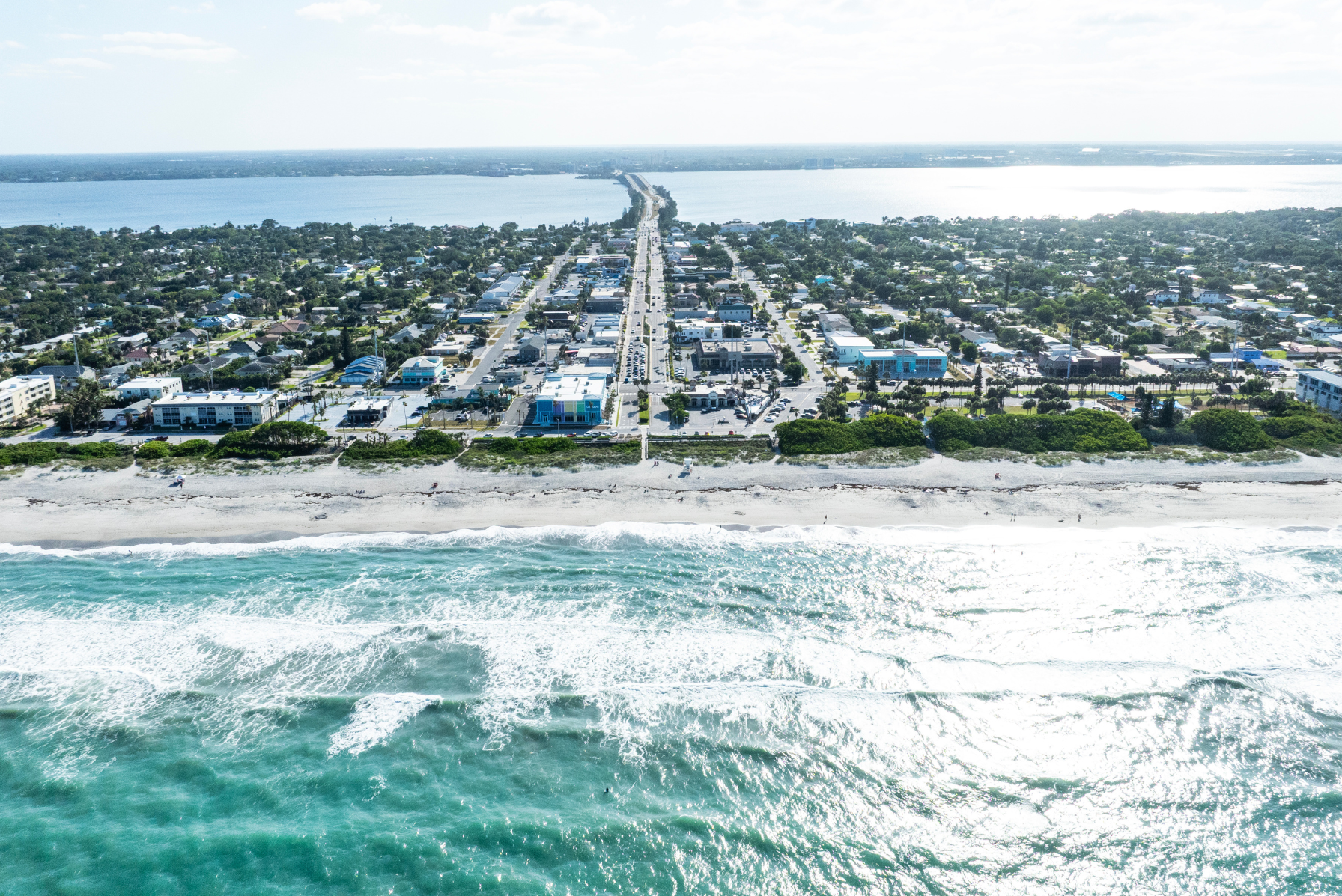 Aerial view of a coastal town with a beachfront and bridge, showcasing clear blue ocean waves, sandy shoreline, and urban layout.