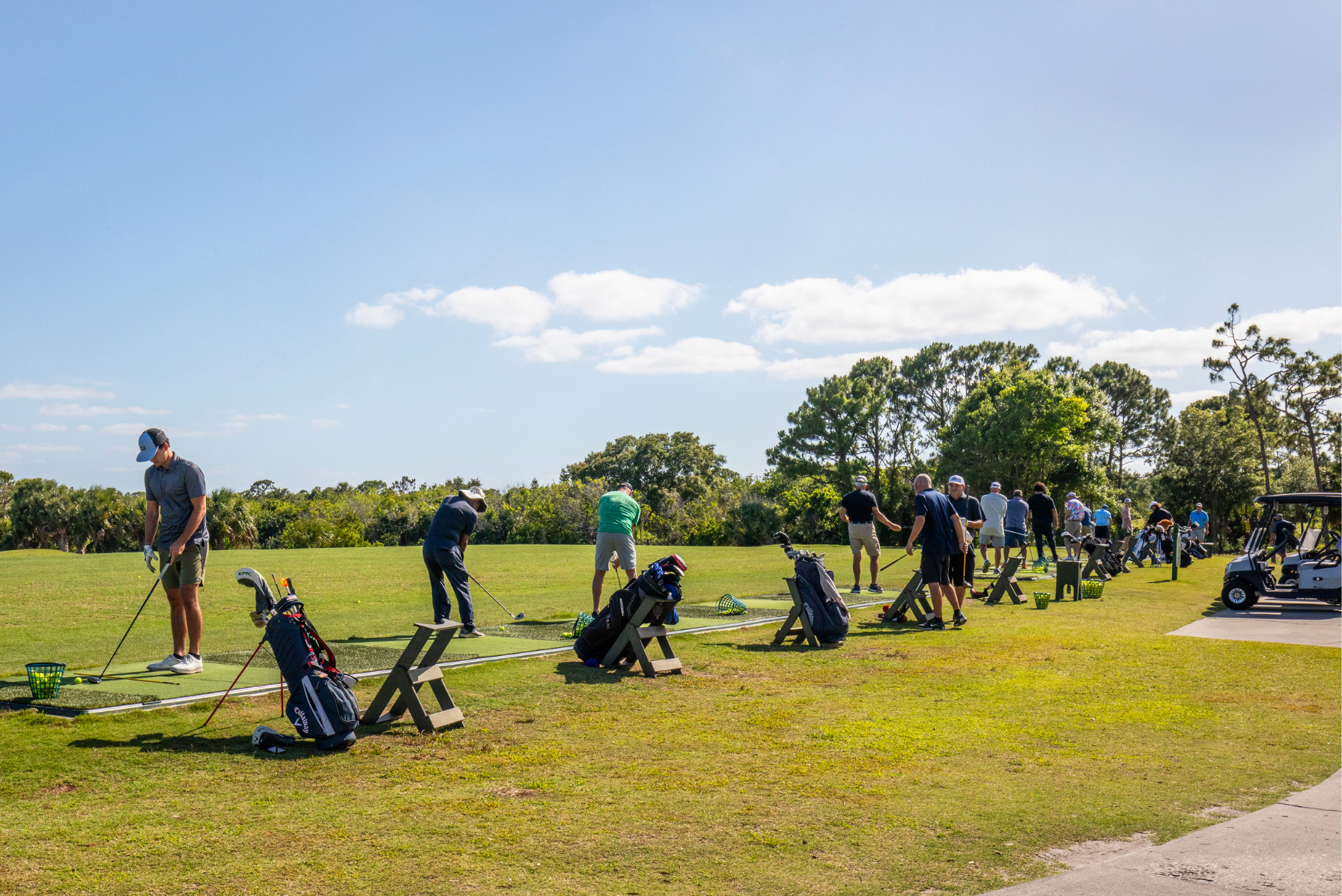 A group of people practicing their swings at a golf driving range on a sunny day, surrounded by green trees and golf bags.