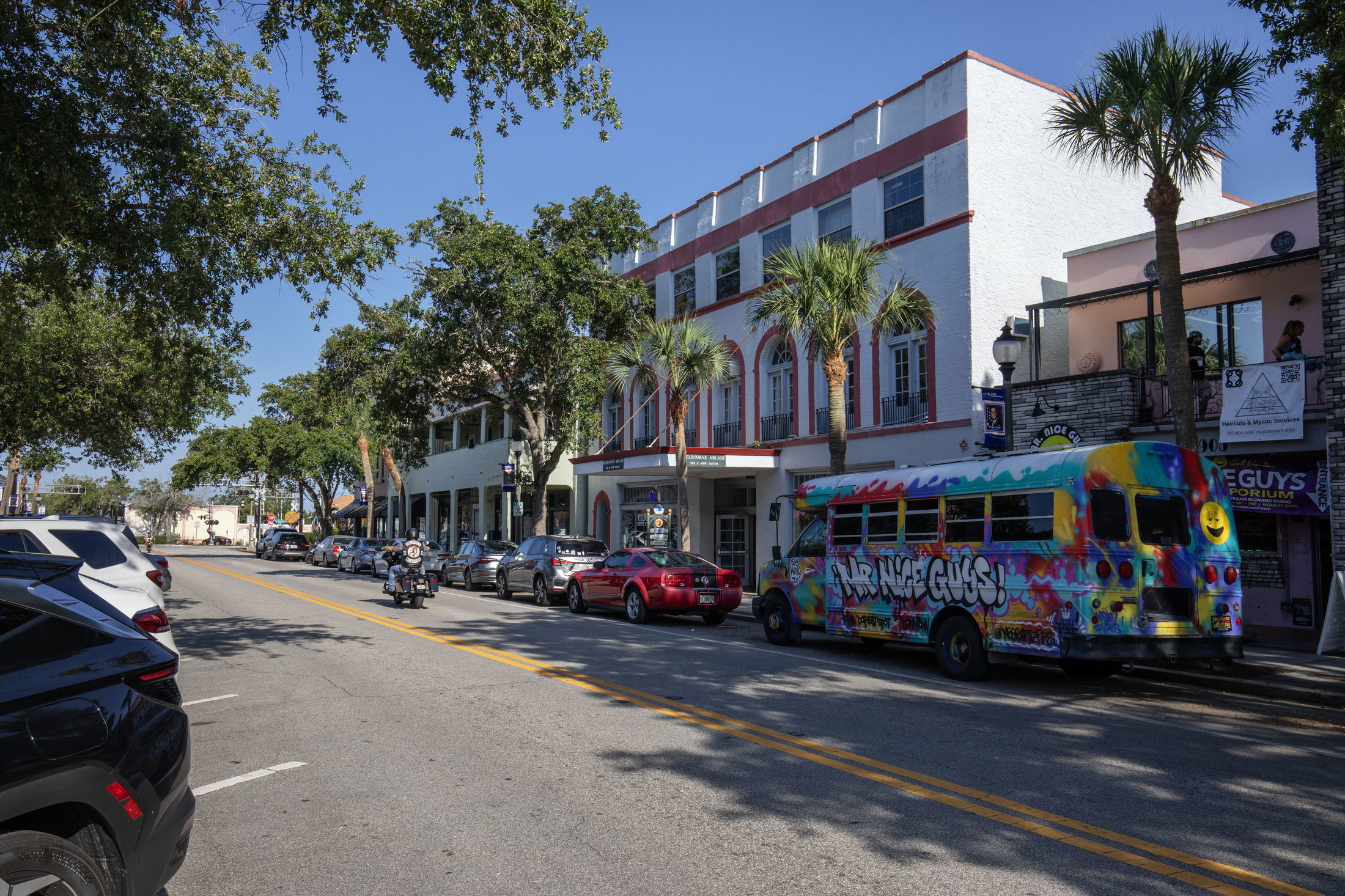 Colorful street scene in a lively town with a vibrant painted bus, parked cars, motorcycles, and palm trees under a clear blue sky.