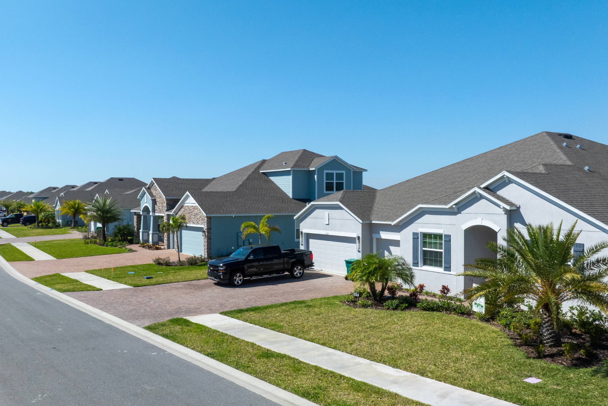 Suburban residential street with modern houses, manicured lawns, and a clear blue sky.