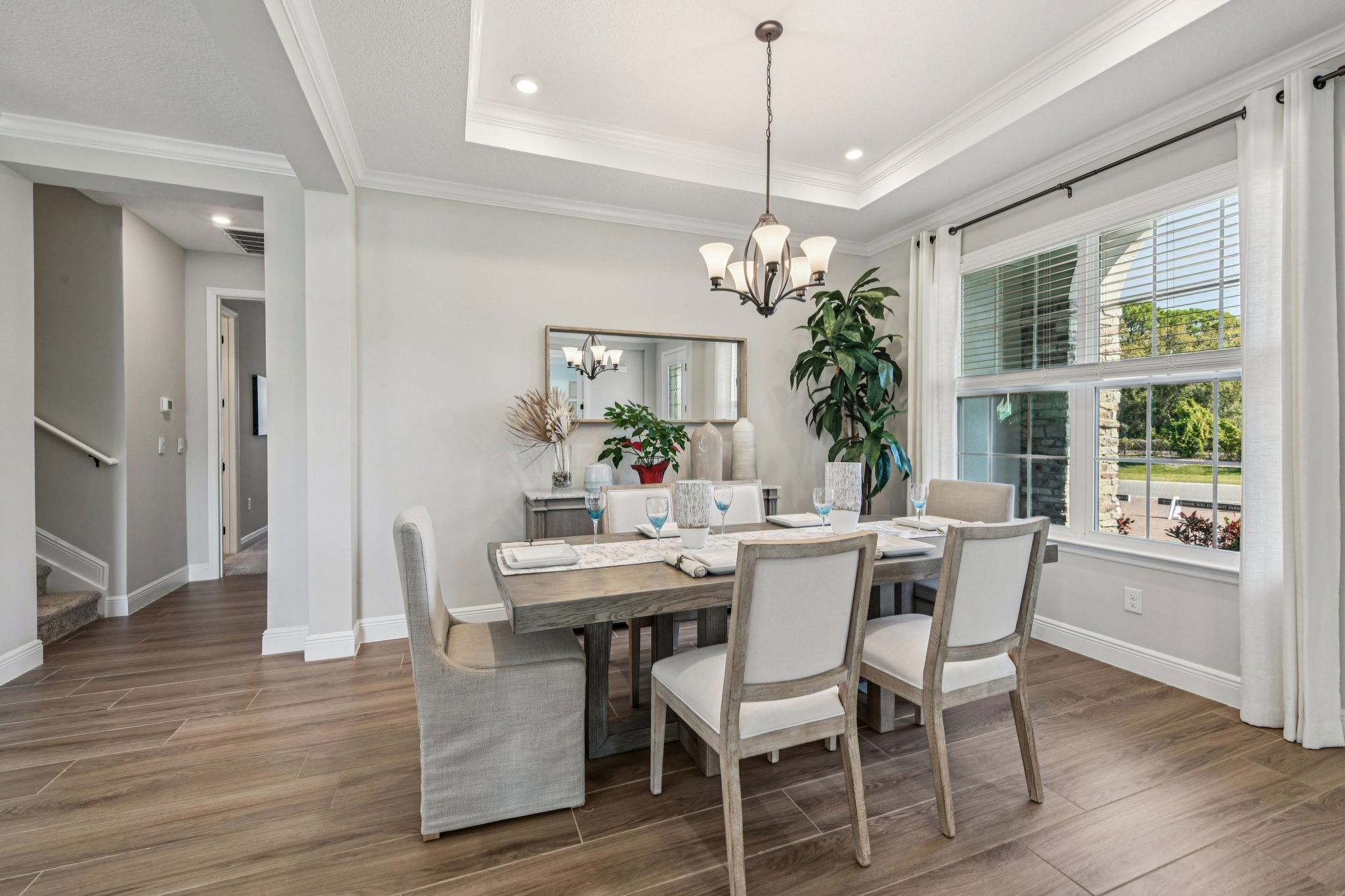 Elegant dining room with a wooden table, cushioned chairs, decorative plants, and a chandelier, featuring large windows and hardwood flooring.