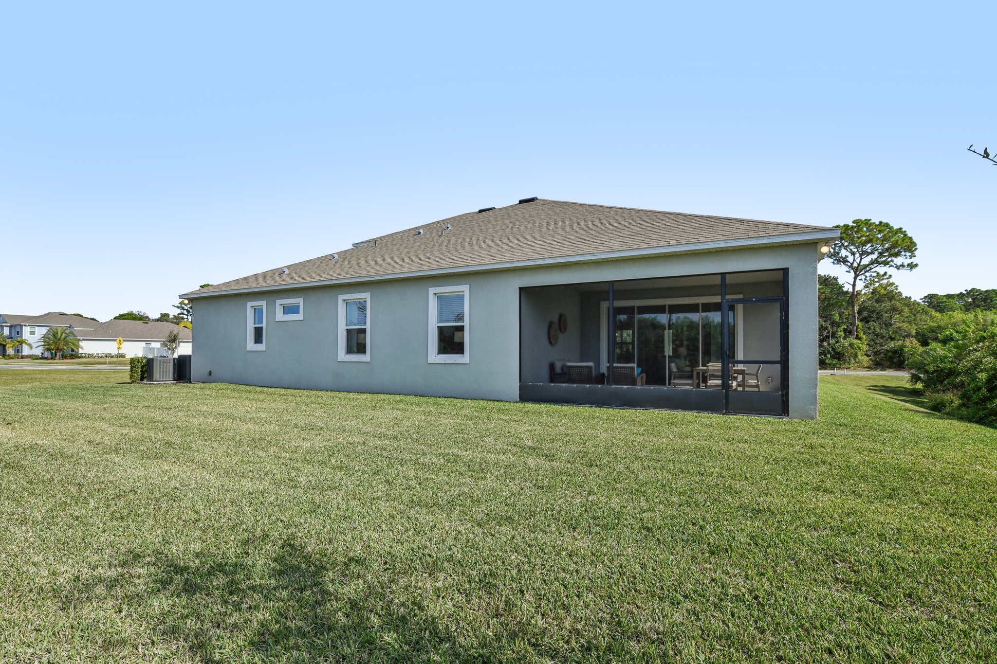 Rear view of a modern, single-story home with a large yard, screened patio, and clean landscaping under a clear blue sky.