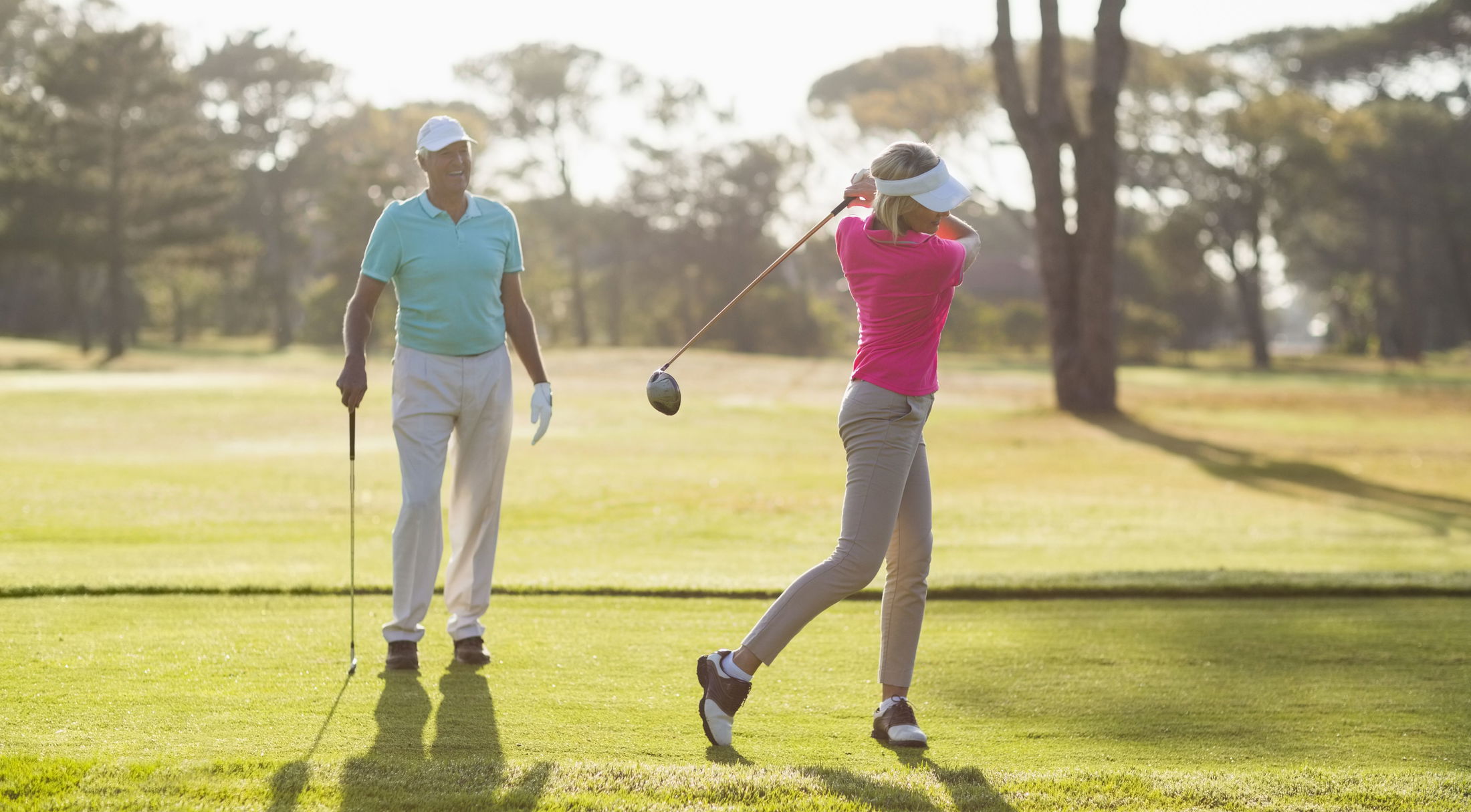 An elderly man watches a woman in a pink top swing a golf club on a sunny golf course.