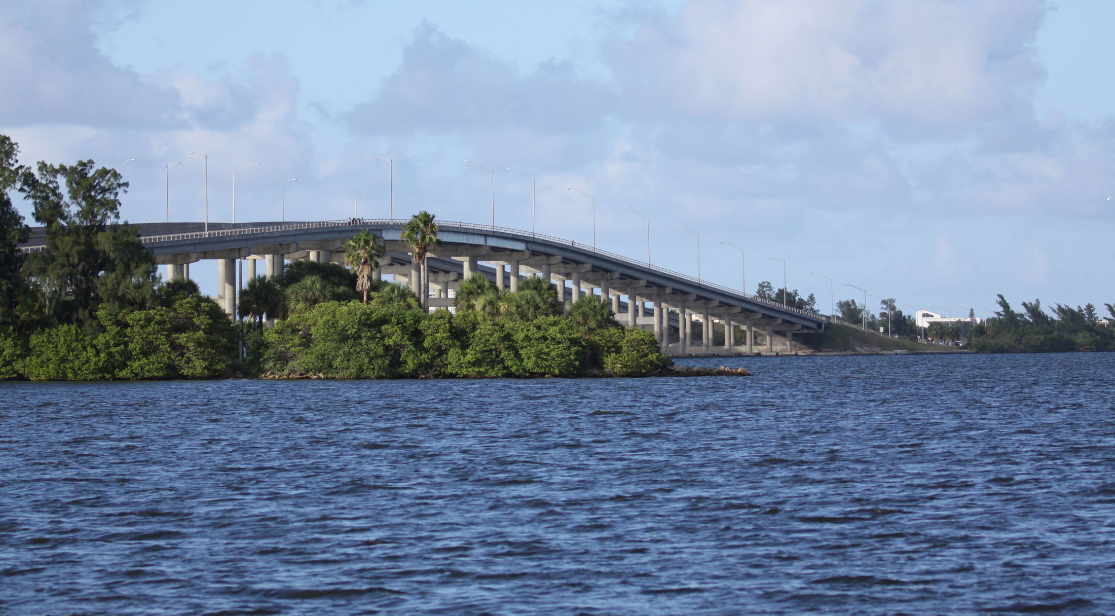 Elevated bridge stretching over a calm blue river with lush green trees beneath, under a partly cloudy sky.