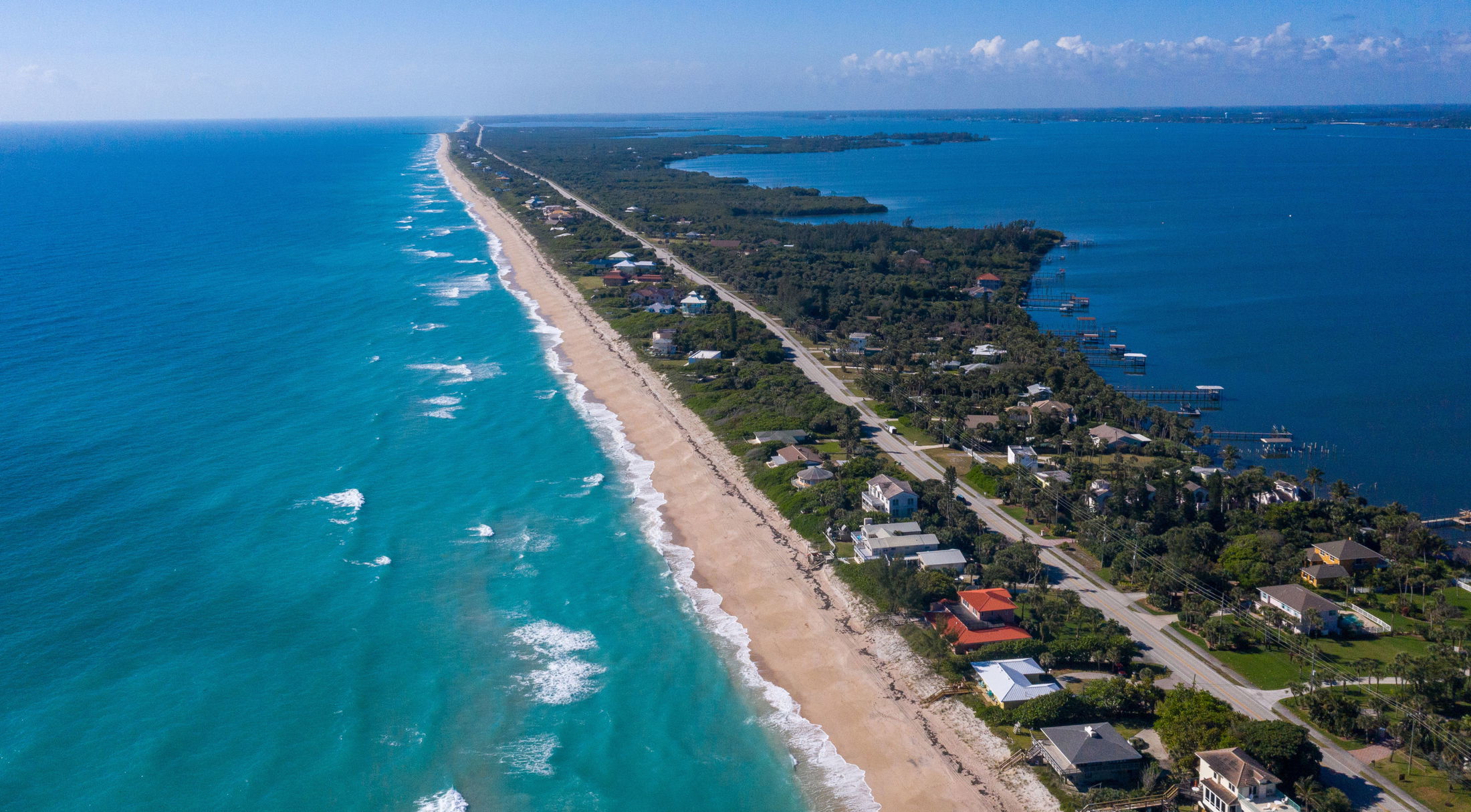 Aerial view of a long, sandy beach with turquoise ocean waves and adjacent residential homes on a narrow strip of land.
