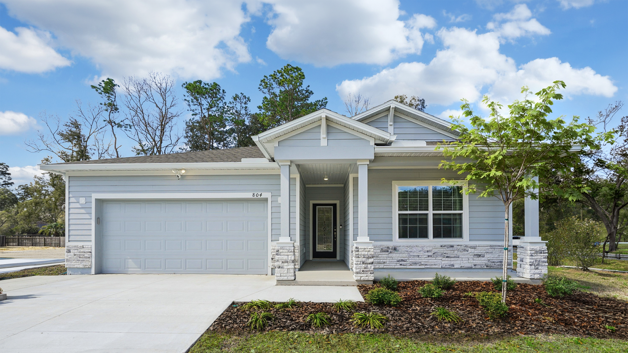 Single-story blue house with stone accents, a front porch, and a two-car garage, surrounded by landscaping and a clear blue sky.