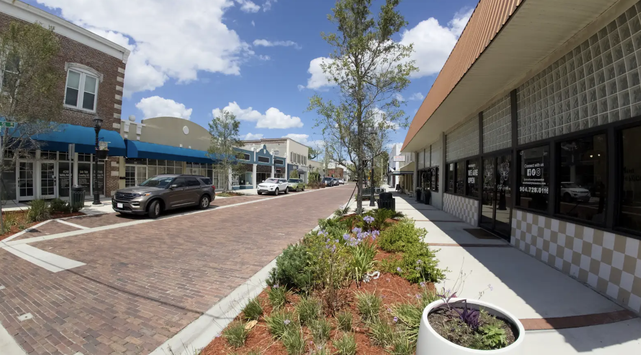 A pedestrian-friendly street featuring brick pavement, flowering plants, and storefronts on a sunny day.