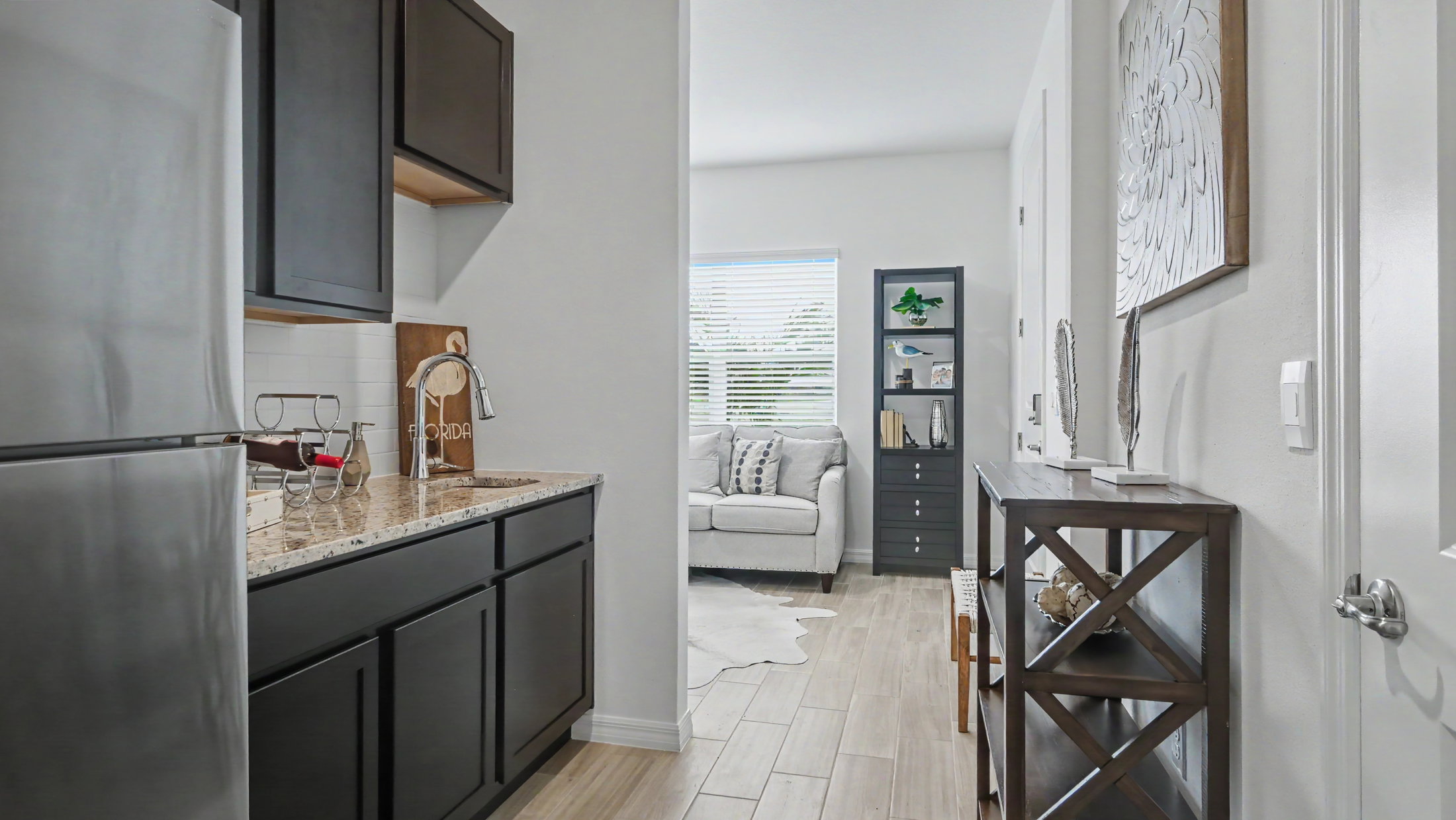 A modern kitchen with dark cabinets and granite countertops leading to a cozy living space with natural light and decorative shelves.