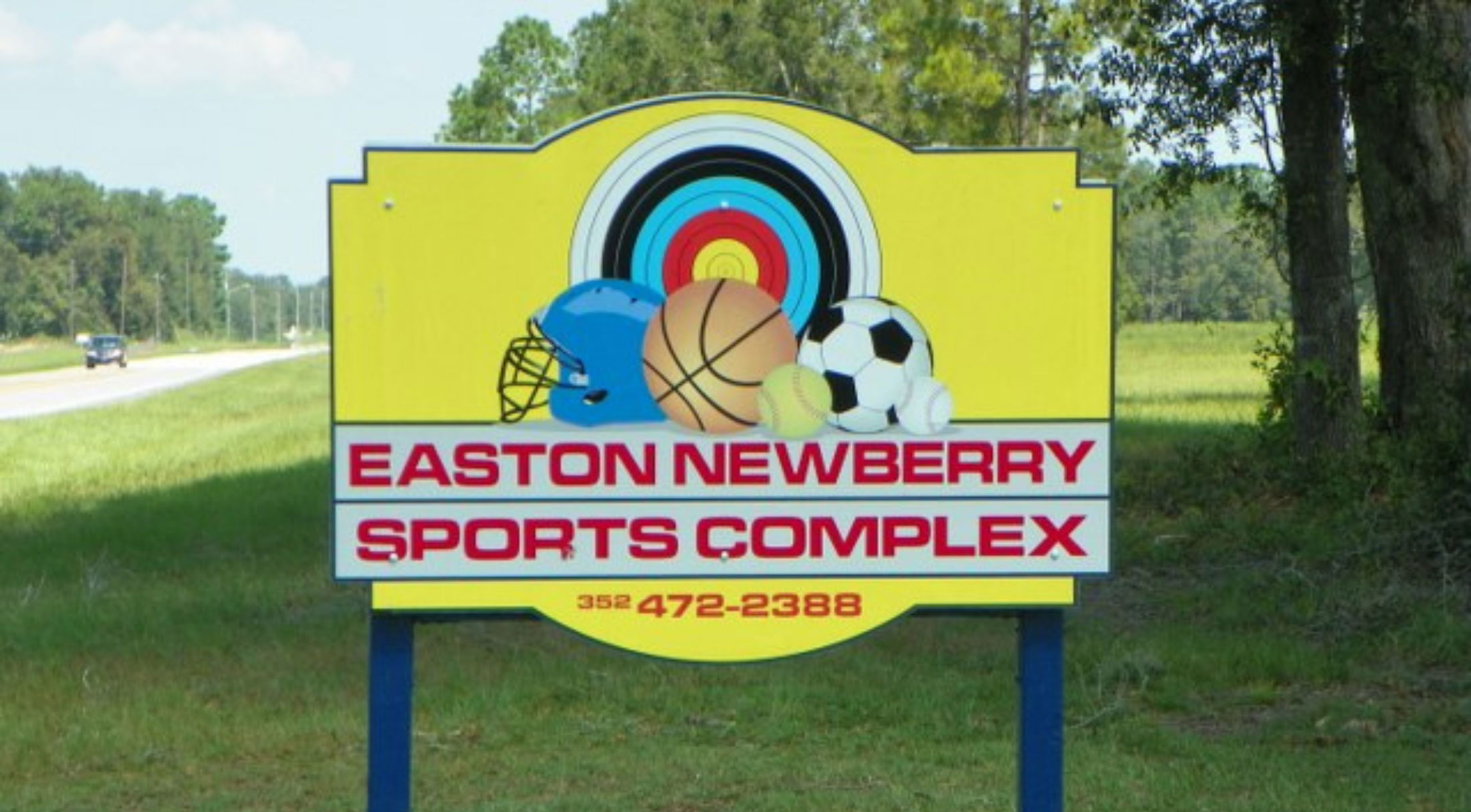 Sign for Easton Newberry Sports Complex featuring helmets, sports balls, and an archery target in a grassy outdoor setting.