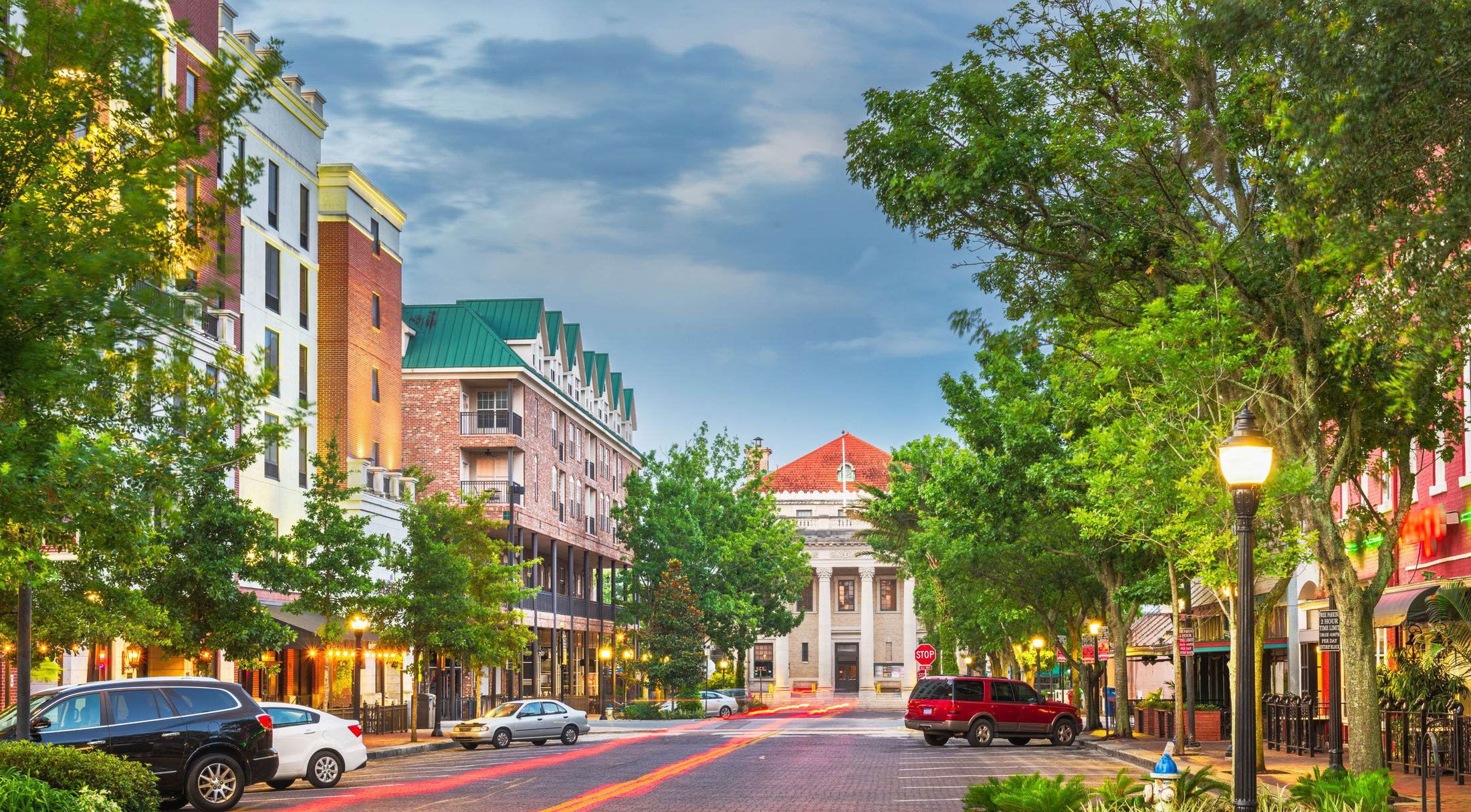 Wide-angle view of a vibrant, tree-lined downtown street in Gainesville, Florida, featuring colorful buildings and historic architecture.