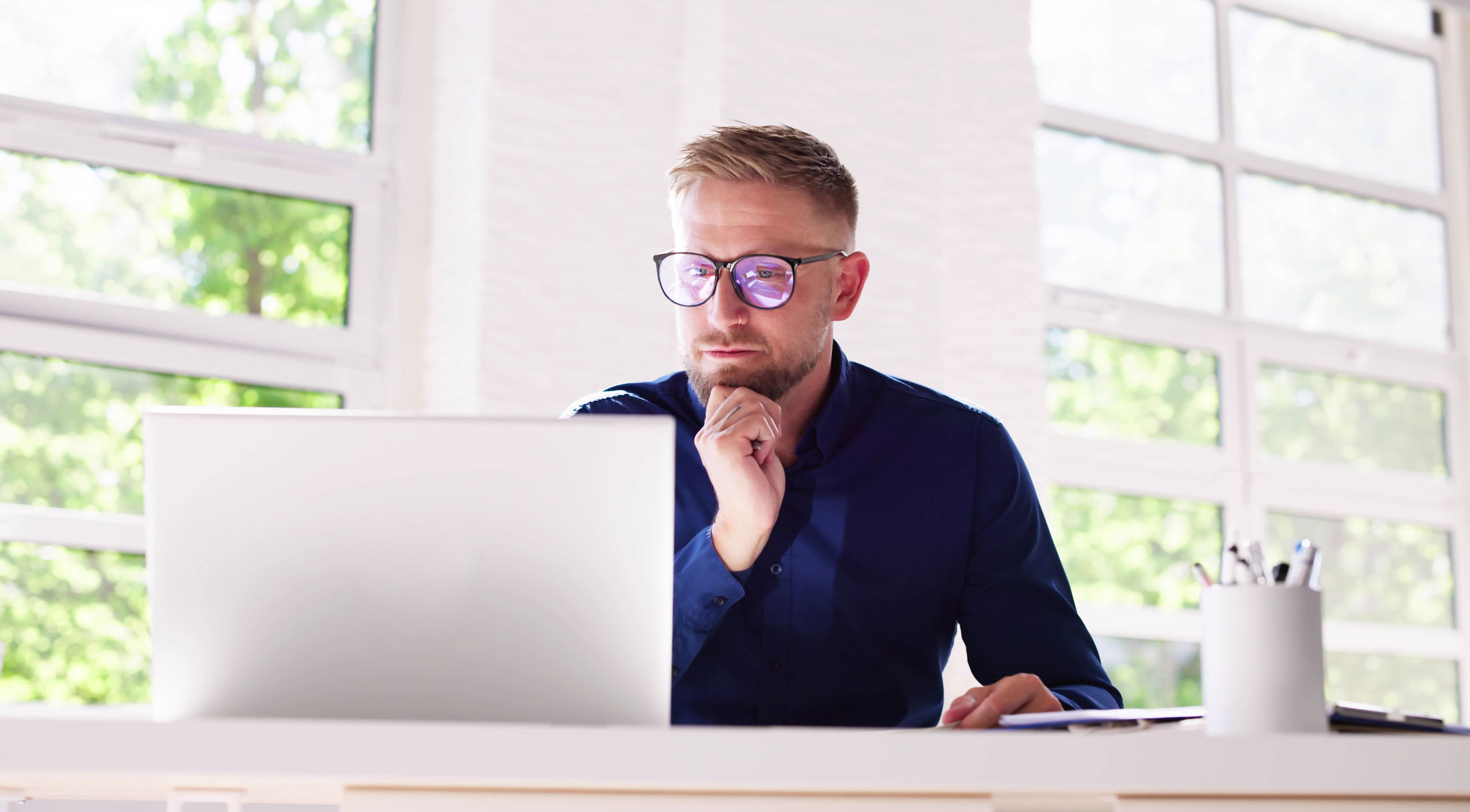 A man in a blue shirt and glasses working on a laptop in a bright office with large windows.