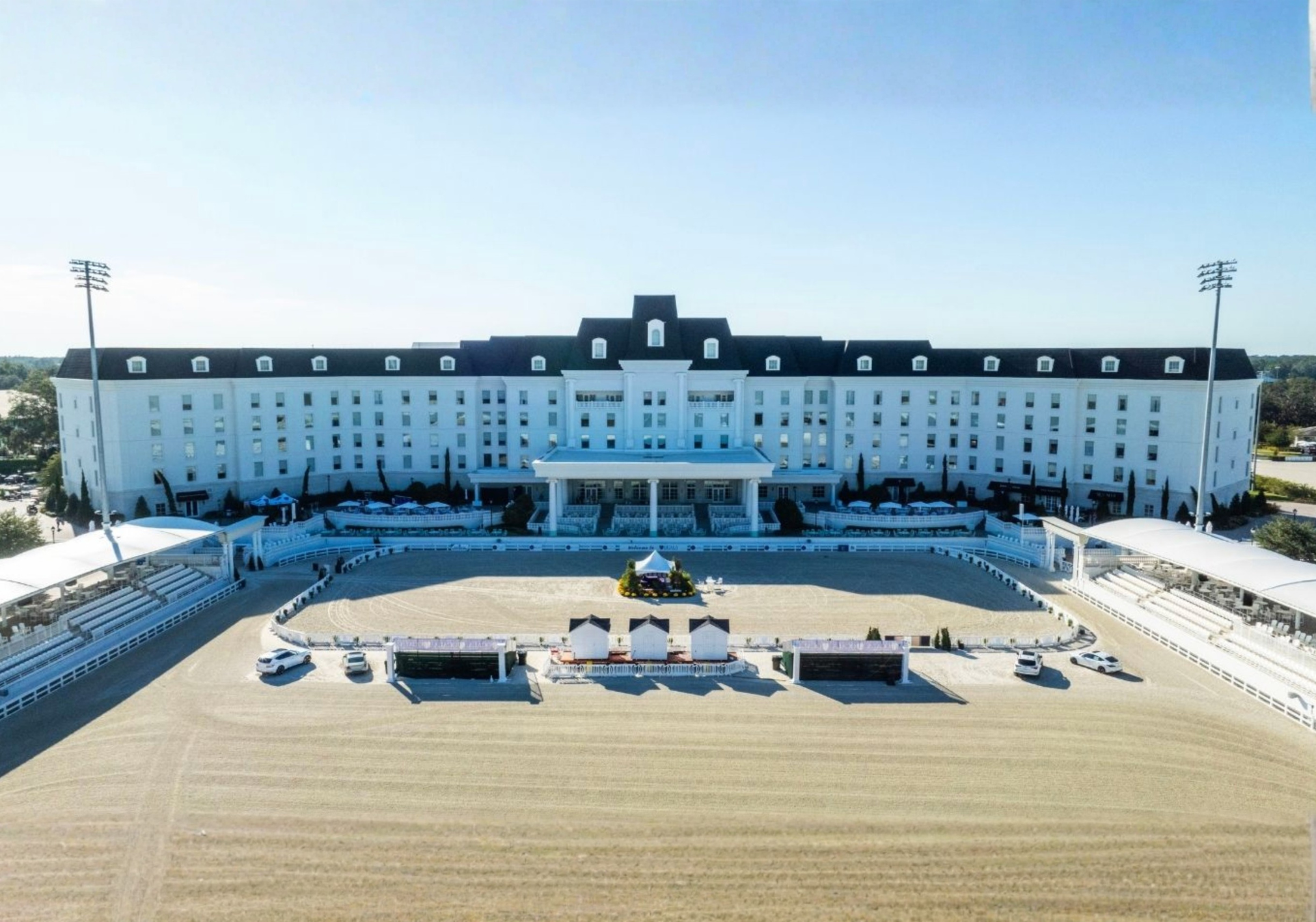 A large white building with multiple stories and a flat roof overlooks an outdoor equestrian arena.