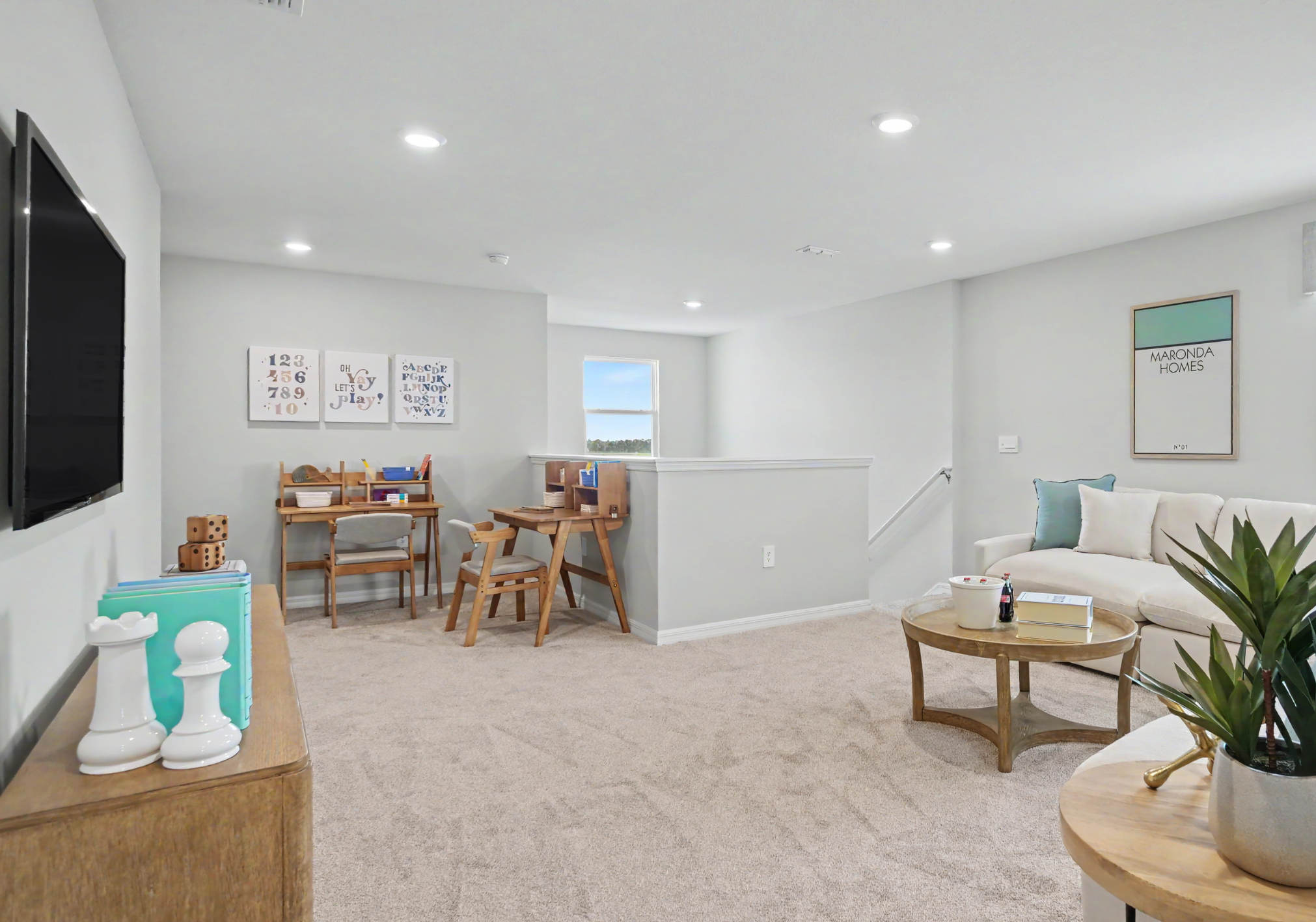 A well-lit living area featuring a beige carpet, a couch with pillows, a wooden coffee table, and two workstations with chairs against a wall adorned with framed art.