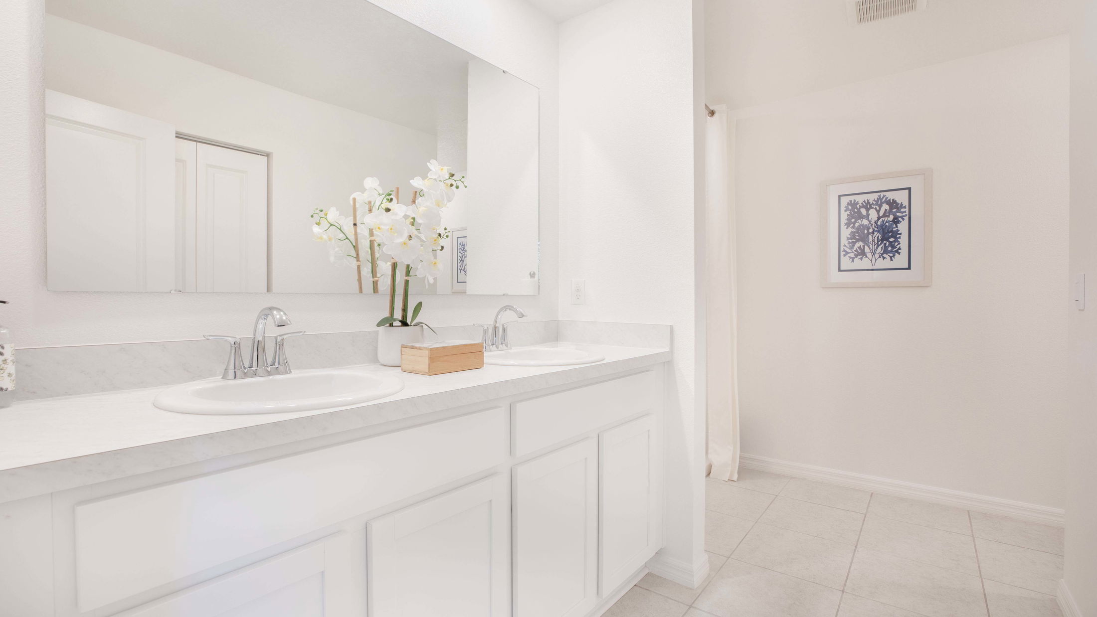 Elegant modern bathroom with dual sinks, white cabinetry, decorative orchid plant, and minimalist wall art.