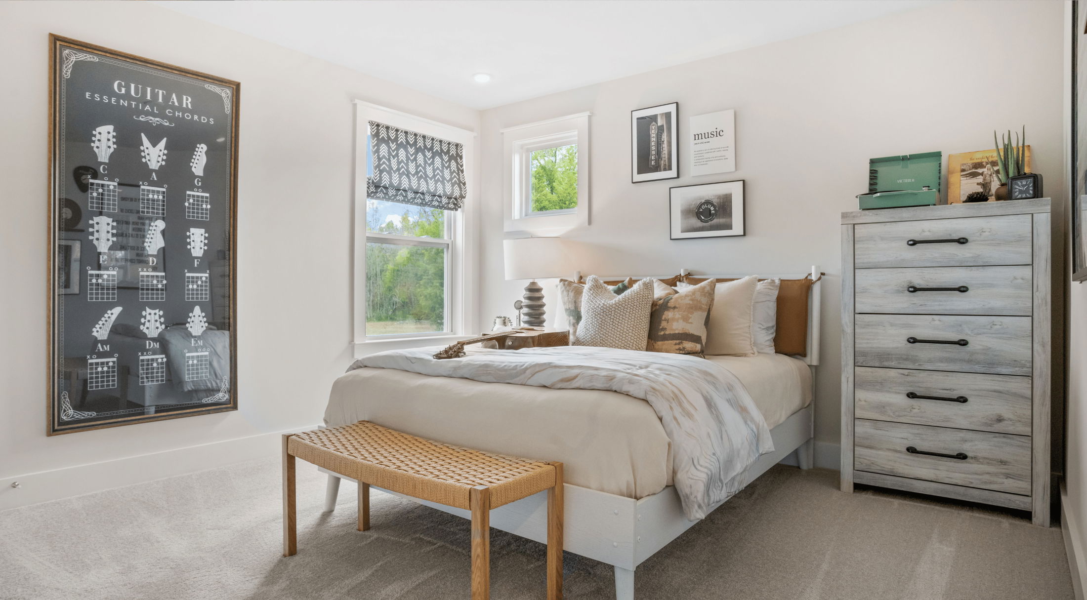 A stylish bedroom featuring a bed with decorative pillows, a wooden bench, a guitar chord chart on the wall, and large windows with natural light.