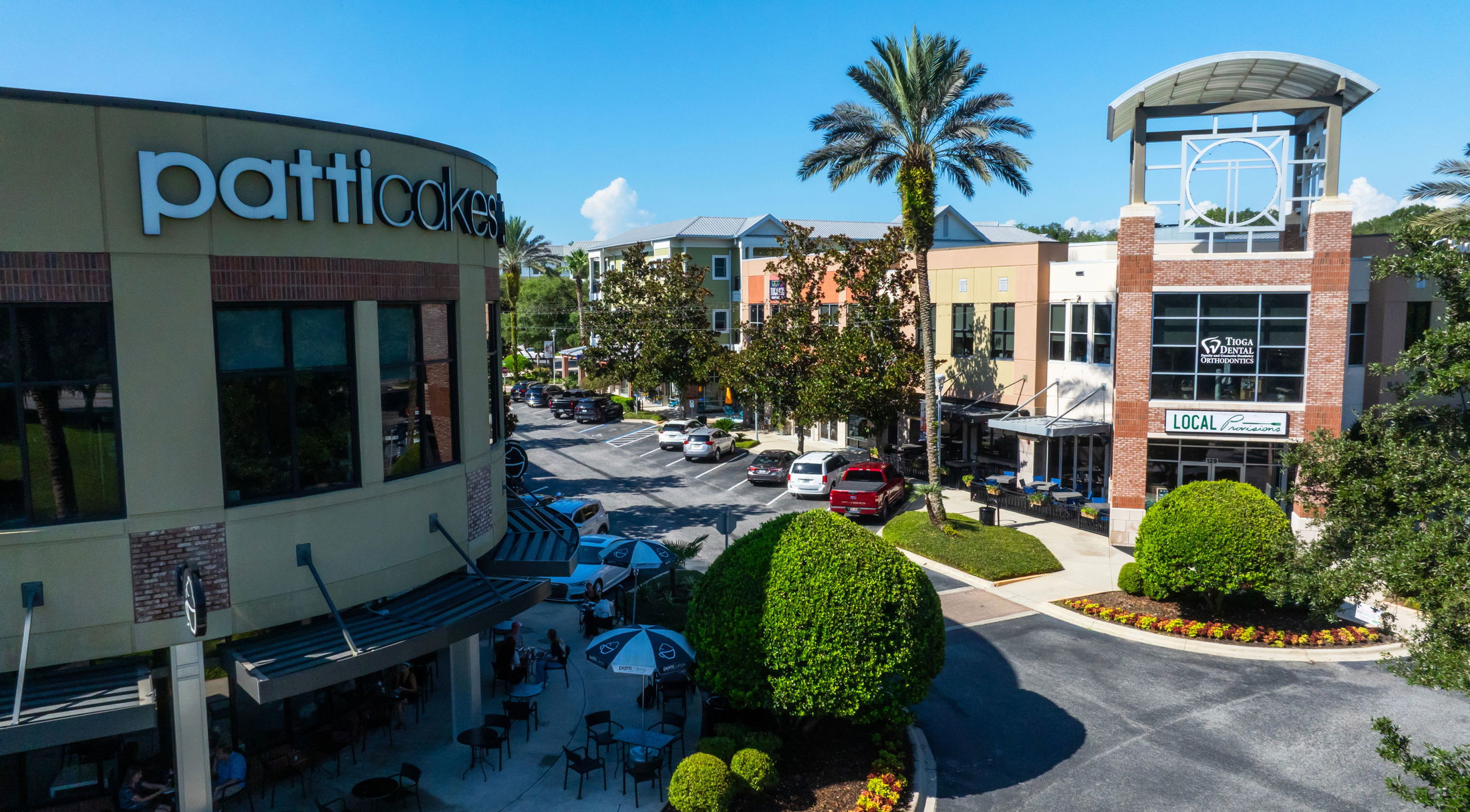 Aerial view of the Patti Cakes bakery and nearby shops in a vibrant shopping center, showcasing palm trees and outdoor seating.