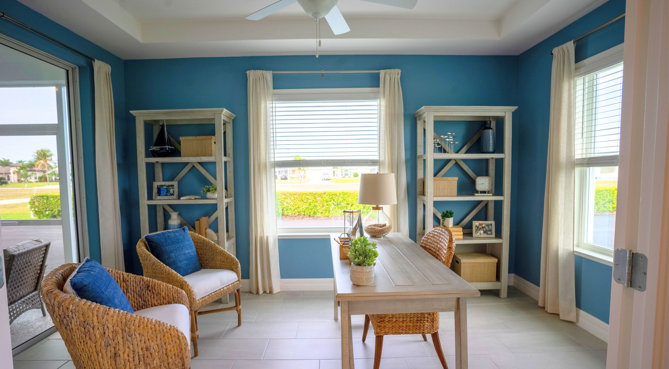A bright and cozy home office featuring blue walls, stylish wicker furniture, and ample natural light from large windows.