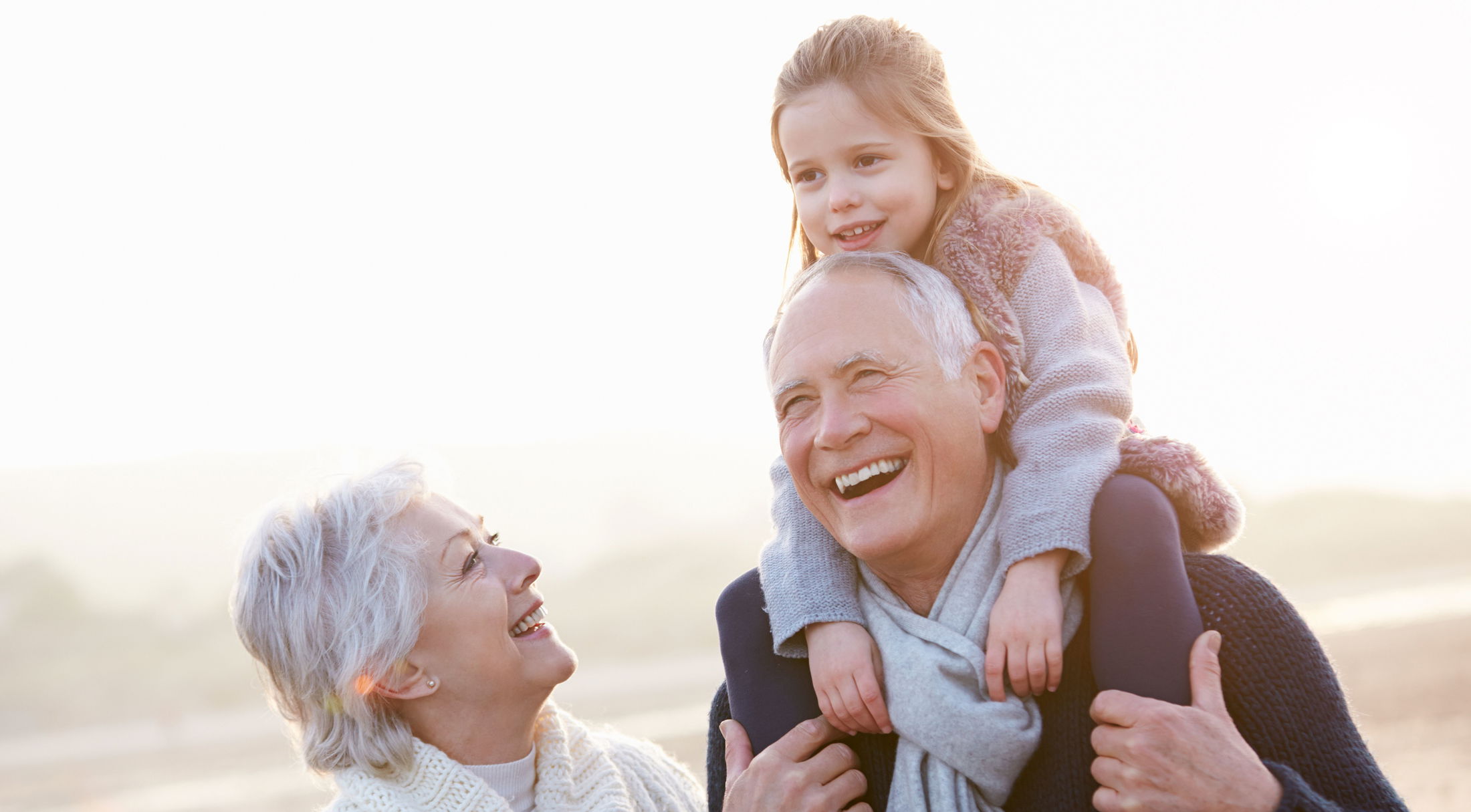 Elderly couple smiling with a young girl on the man\\\'s shoulders enjoying a sunny day outdoors.