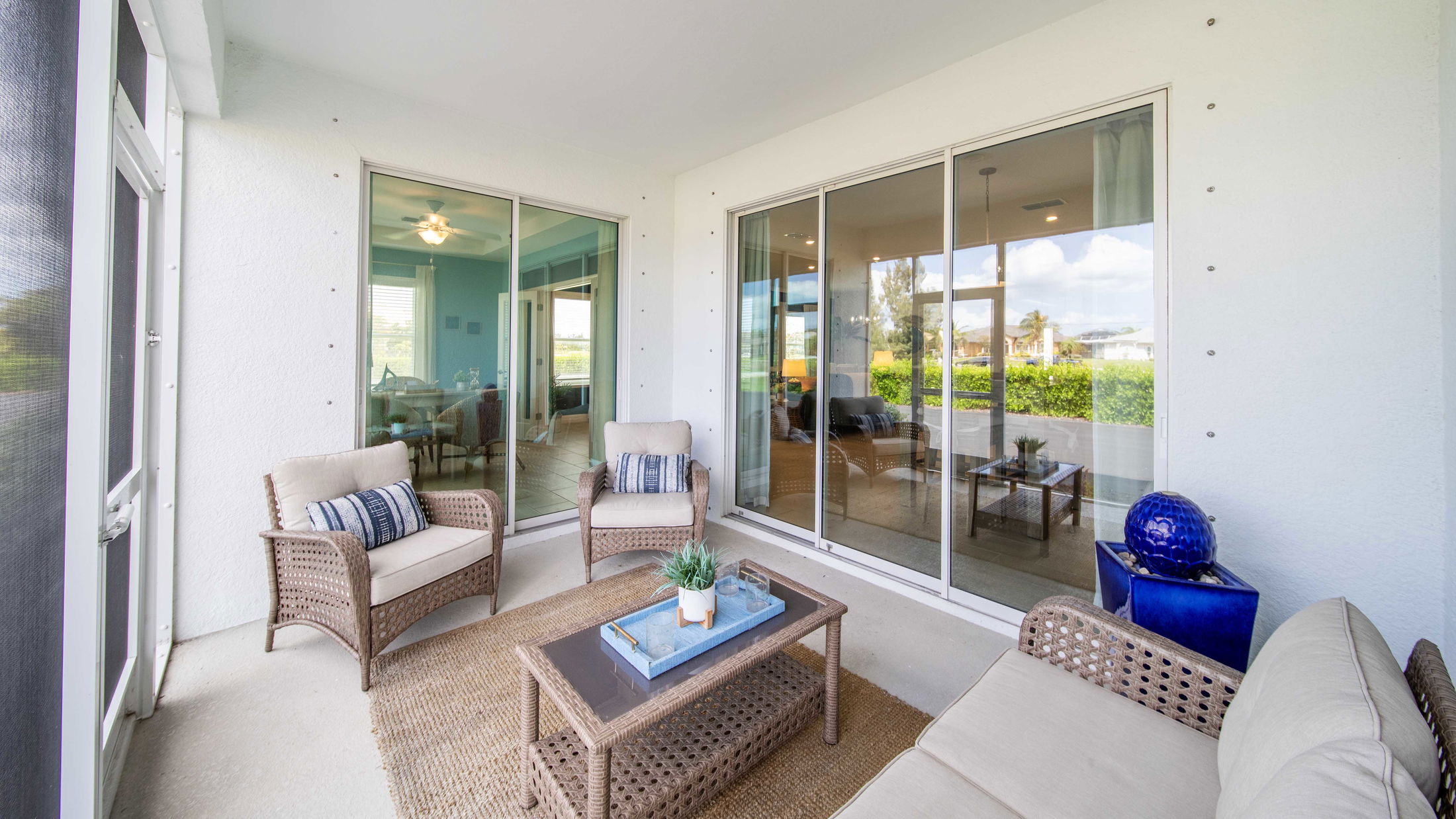 Covered patio with wicker furniture and glass table, overlooking lush greenery through large sliding glass doors.