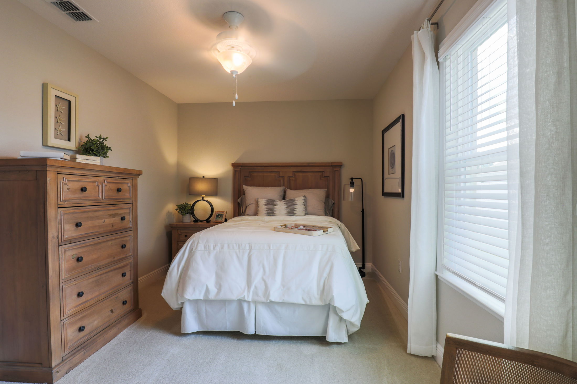 Cozy bedroom with wooden furniture, including a bed, nightstand, and dresser, accented by soft lighting and a large window with white blinds.