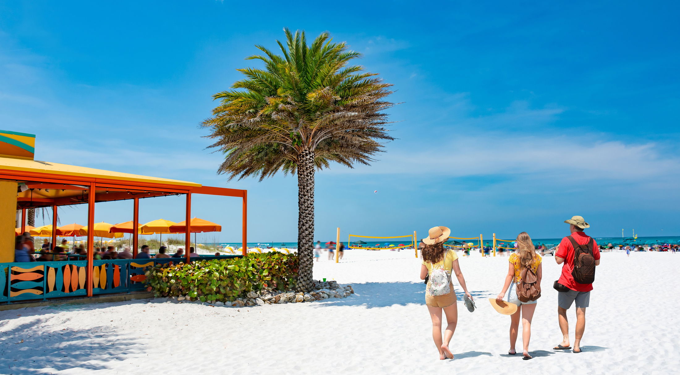 Three people walking on a sandy beach near a colorful beachside cafe and a palm tree on a sunny day with bright blue skies.